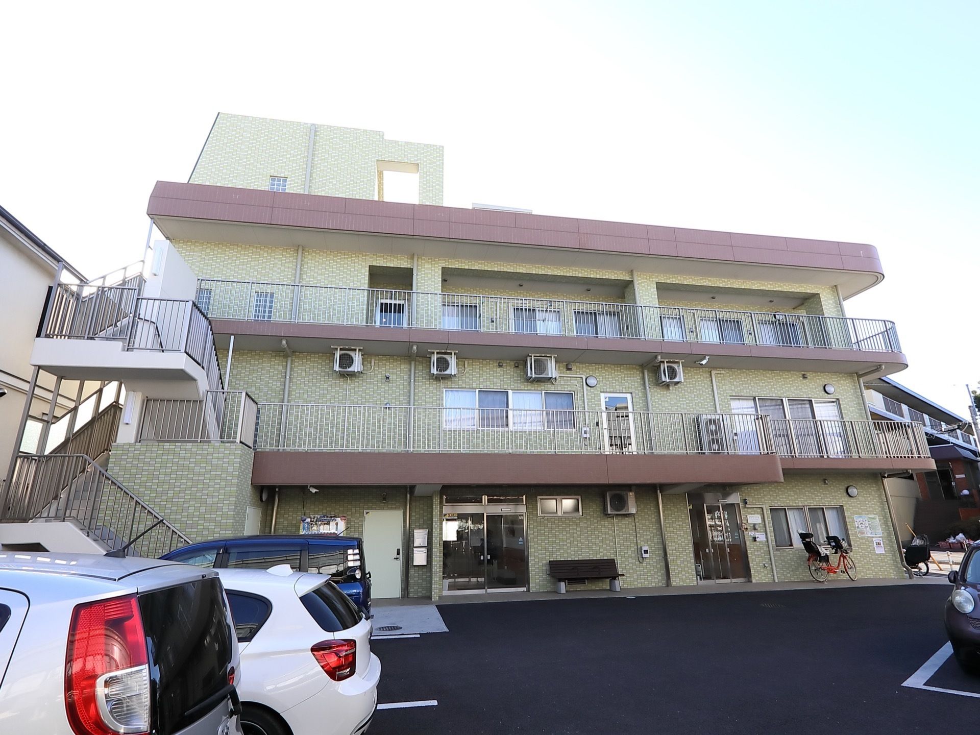 ひさしの部分はあずき色で、外壁は淡い草色の３階建ての建物である。左端に外階段が設置されている。建物の前面が駐車場となっている。