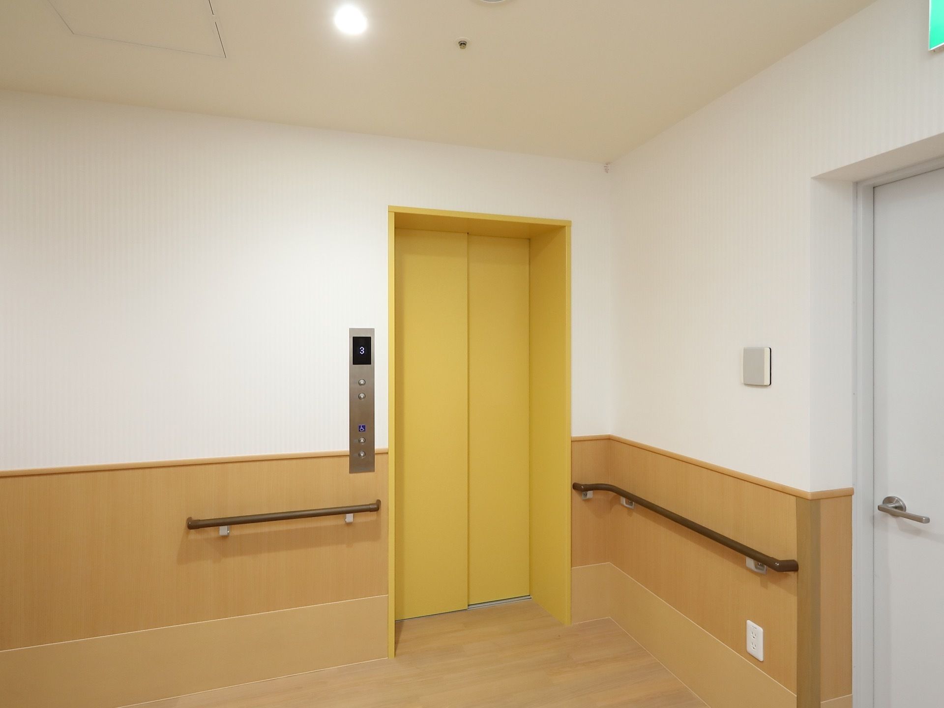 通路の端に設置されたエレベーターである。ドアは黄色に塗られている。エレベーター横の壁には腰高の位置に手すりが設置されている。