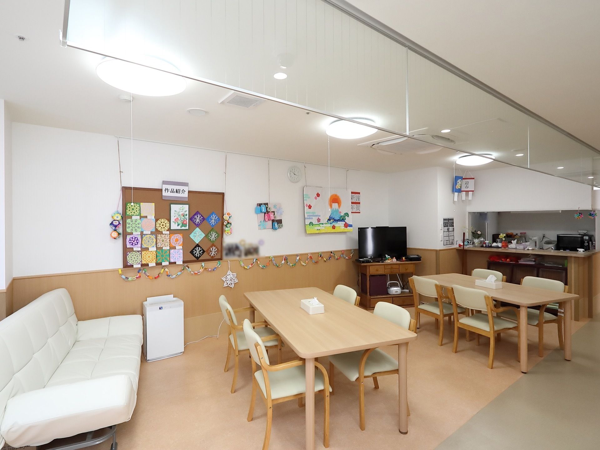 左端に白いソファーがある。木製のダイニングテーブルと椅子がセットされ、２組置かれている。奥に厨房があり、壁際にテレビが設置されている。