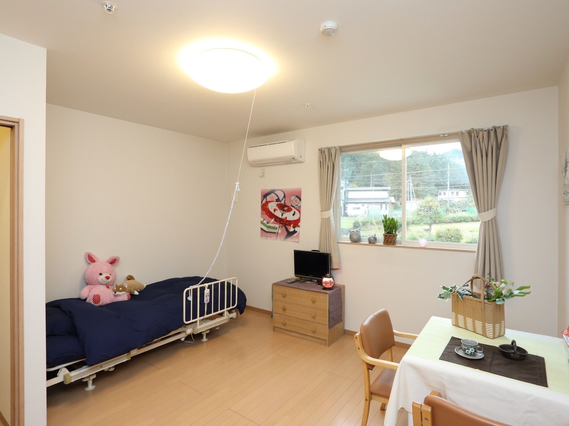 壁側に青い布団が敷かれたベッドがあり、その上にぬいぐるみが置かれている。窓には植物や小物が並べて飾られている。
