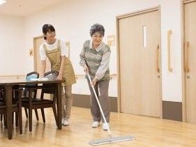 モップで床掃除をしている入居者をやさしく見守るスタッフの様子。
