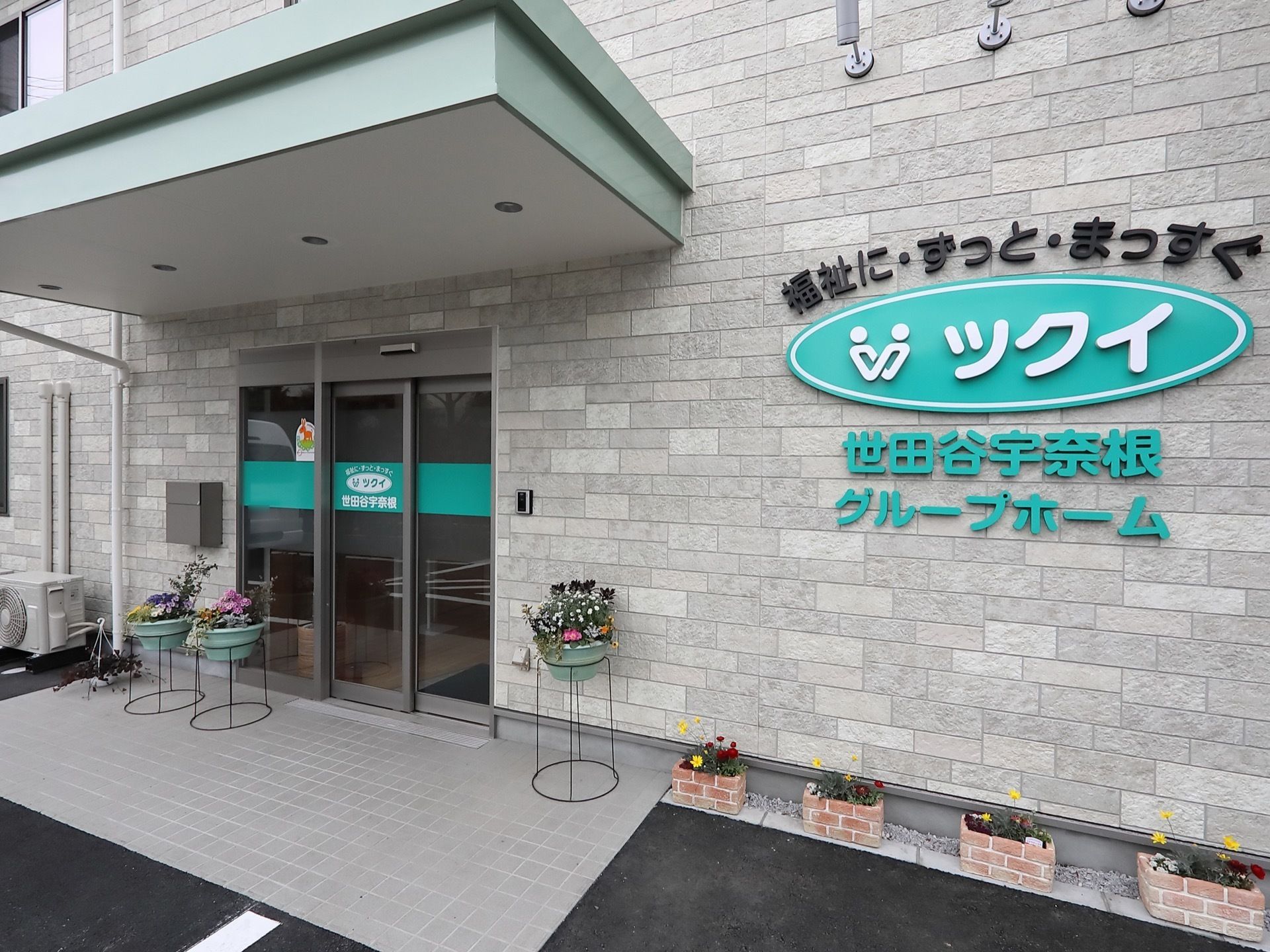 ひさしが設けられたエントランスである。壁際に花が植えられたプランターが並べられている。インターホンと郵便受けが壁に設置されている。