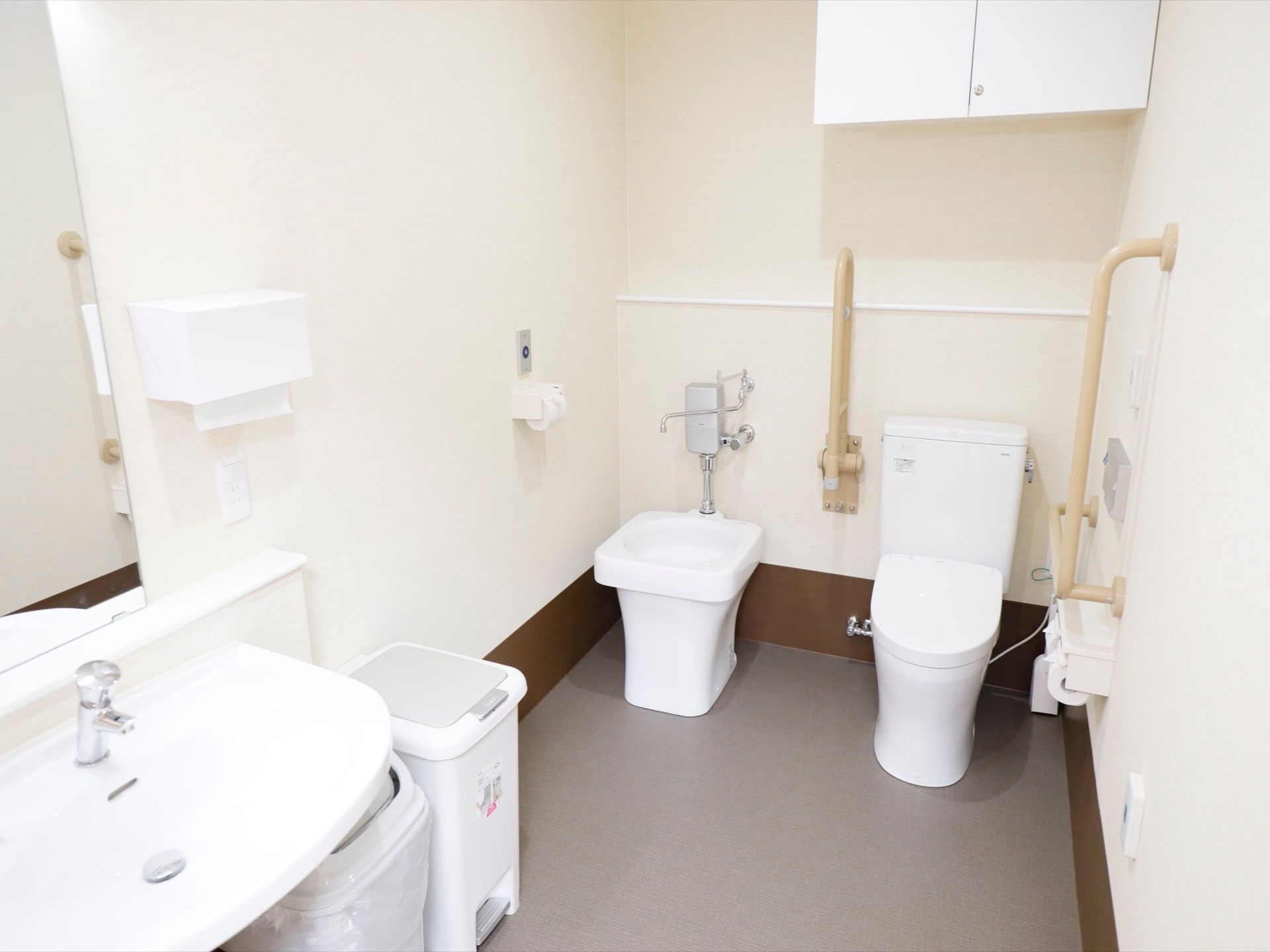 広々とした清潔感のあるオストメイト対応トイレである。手すりや洗面スペース、ゴミ箱、ナースコールが完備されている。