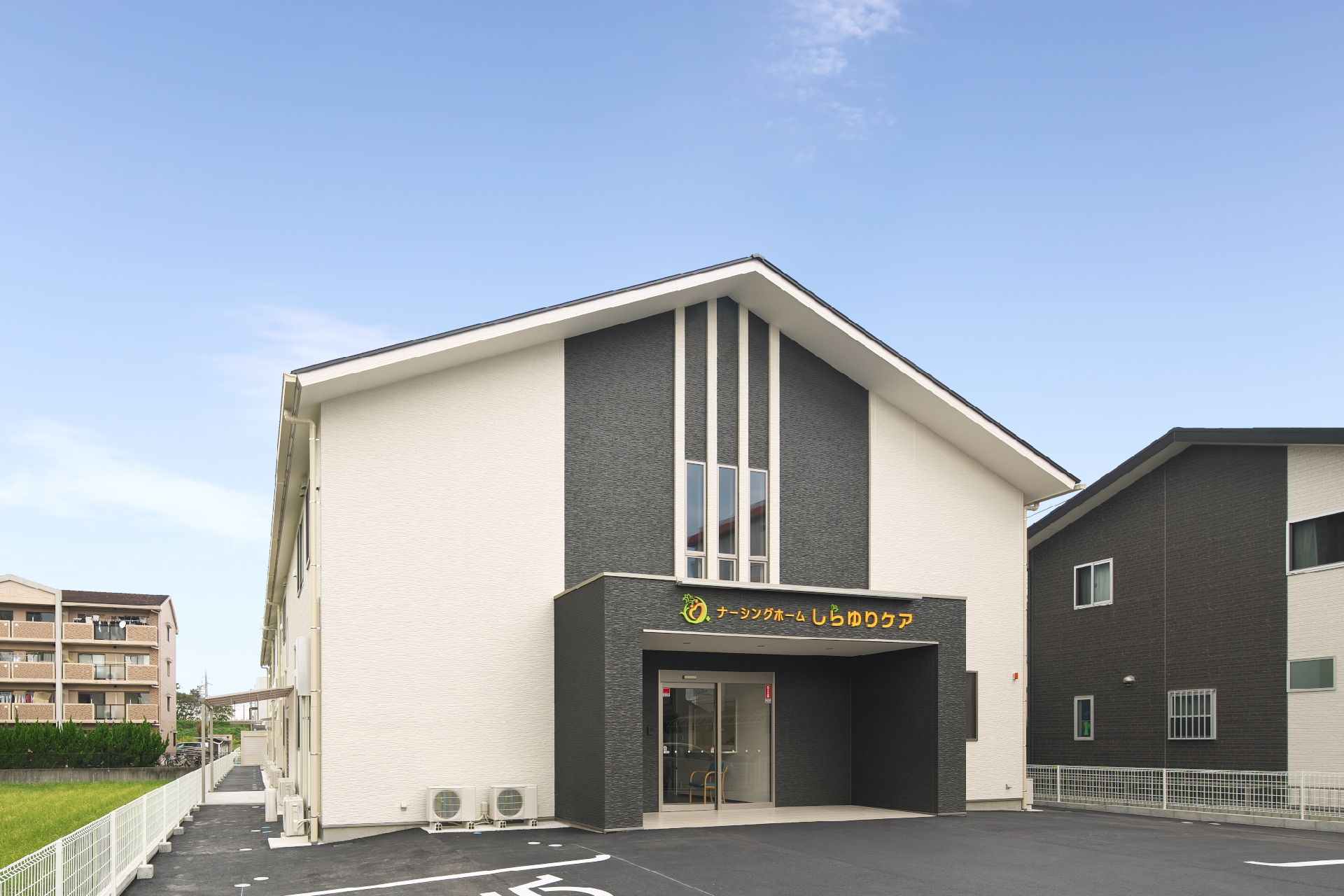 2階建ての建物。白い外壁にグレーのラインが入っていて屋根はブラック。敷地内には広い駐車スペースがある。