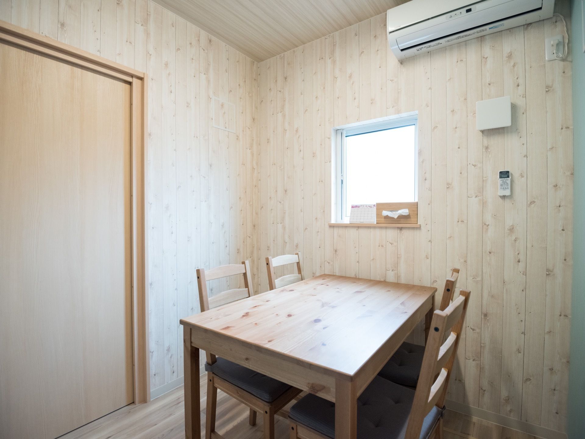 扉や壁、天井、テーブルなど全て木目調で統一されたフローリングの部屋がある。窓があり、その上にはエアコンが設置されている。