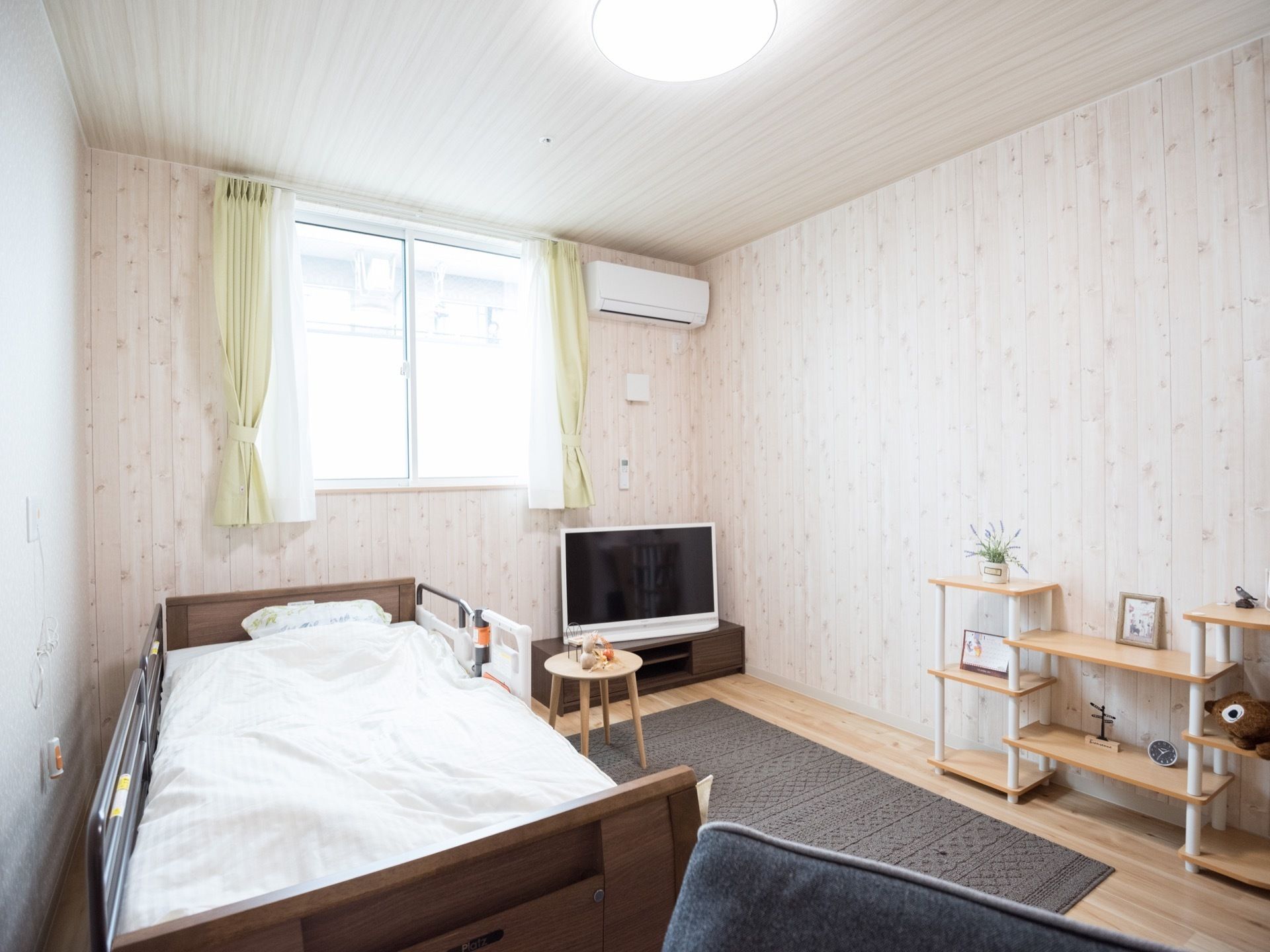 フローリングの部屋に電動ベッドが置かれている。部屋の隅にはテレビ台があり、窓の横にはエアコンが設置されている。