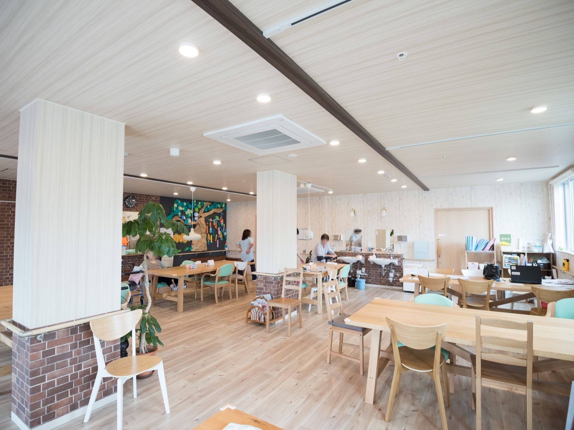 木材の温かみがあるカフェ風の食堂で、スタッフが２人作業をしている。空調が付いていて、奥の壁際には洗面設備がある。