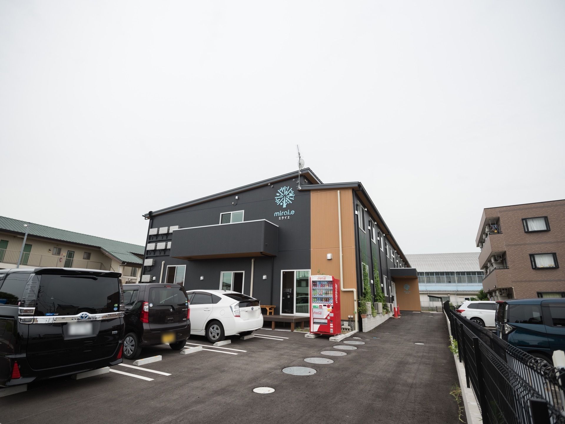 紺色とオレンジ色の外壁が印象的な２階建ての建物がある。手前には舗装された駐車場があり、車が３台停められている。