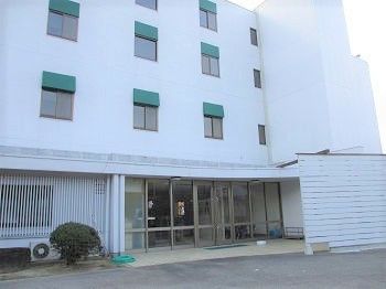 白を基調とした４階建ての建物。玄関はガラスの自動ドアで、玄関横には木が植えられている。