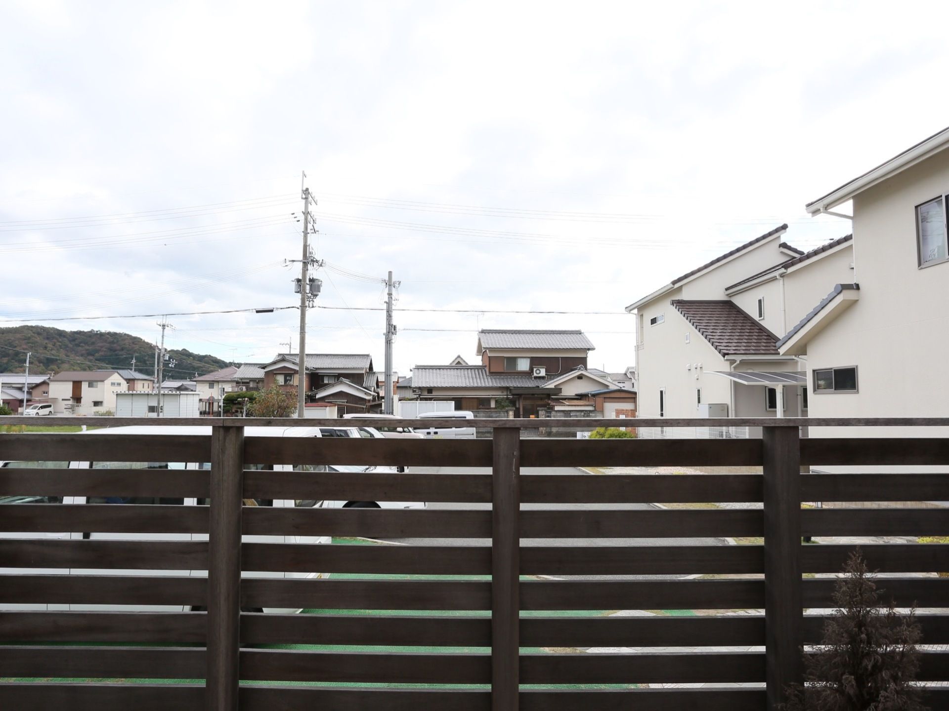 建物の前には木のフェンスが立てられており、駐車場に車が停められているのが見える。奥には住宅街が広がる。