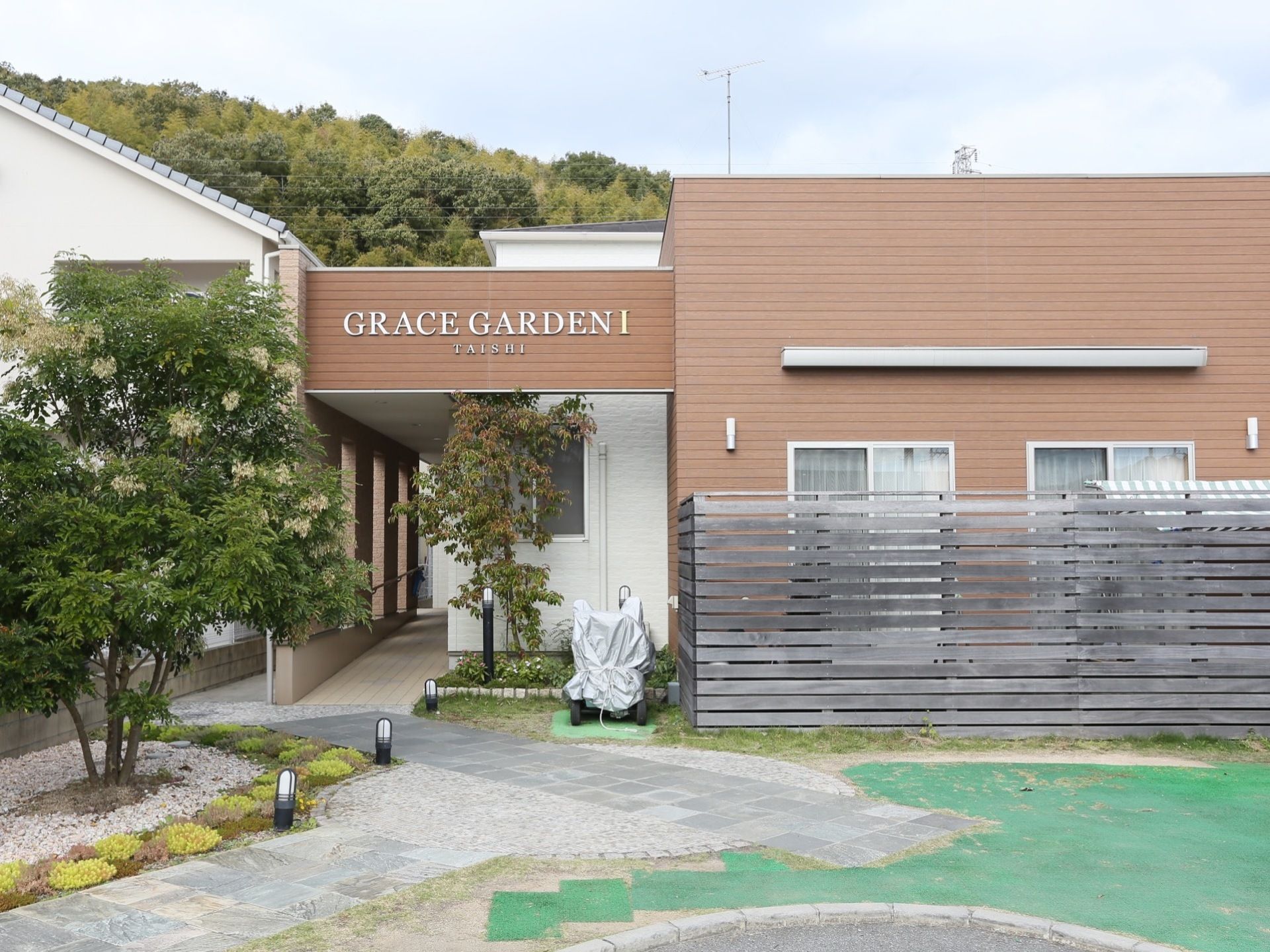 スタイリッシュな外観の建物で、庭に木が植えられている。施設入口は屋根がついており、スロープが設置されている。