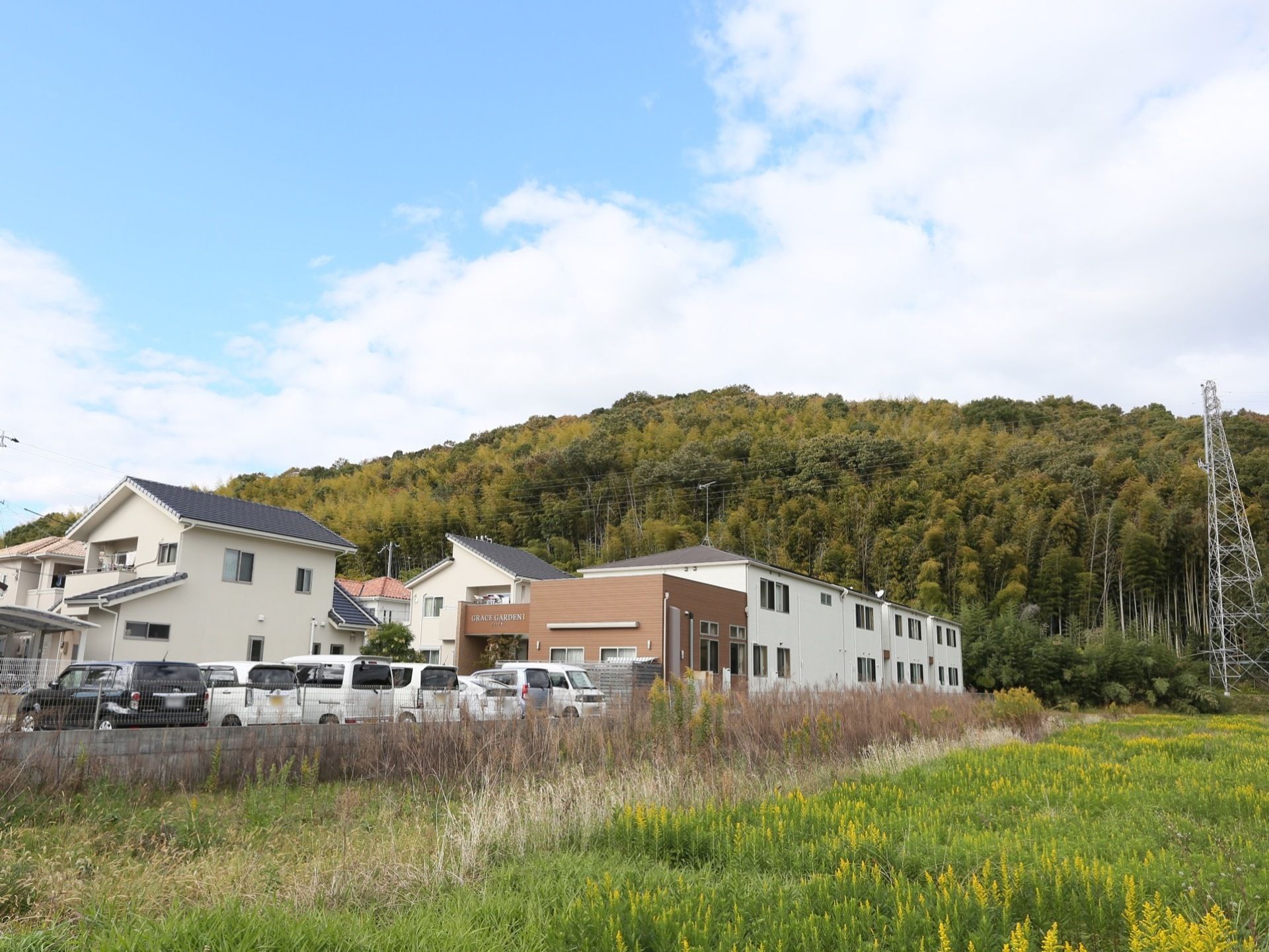 緑豊かな場所に建てられたグループホームで、敷地内に広い駐車場を完備している。後ろには山があり、横には原っぱが広がる。