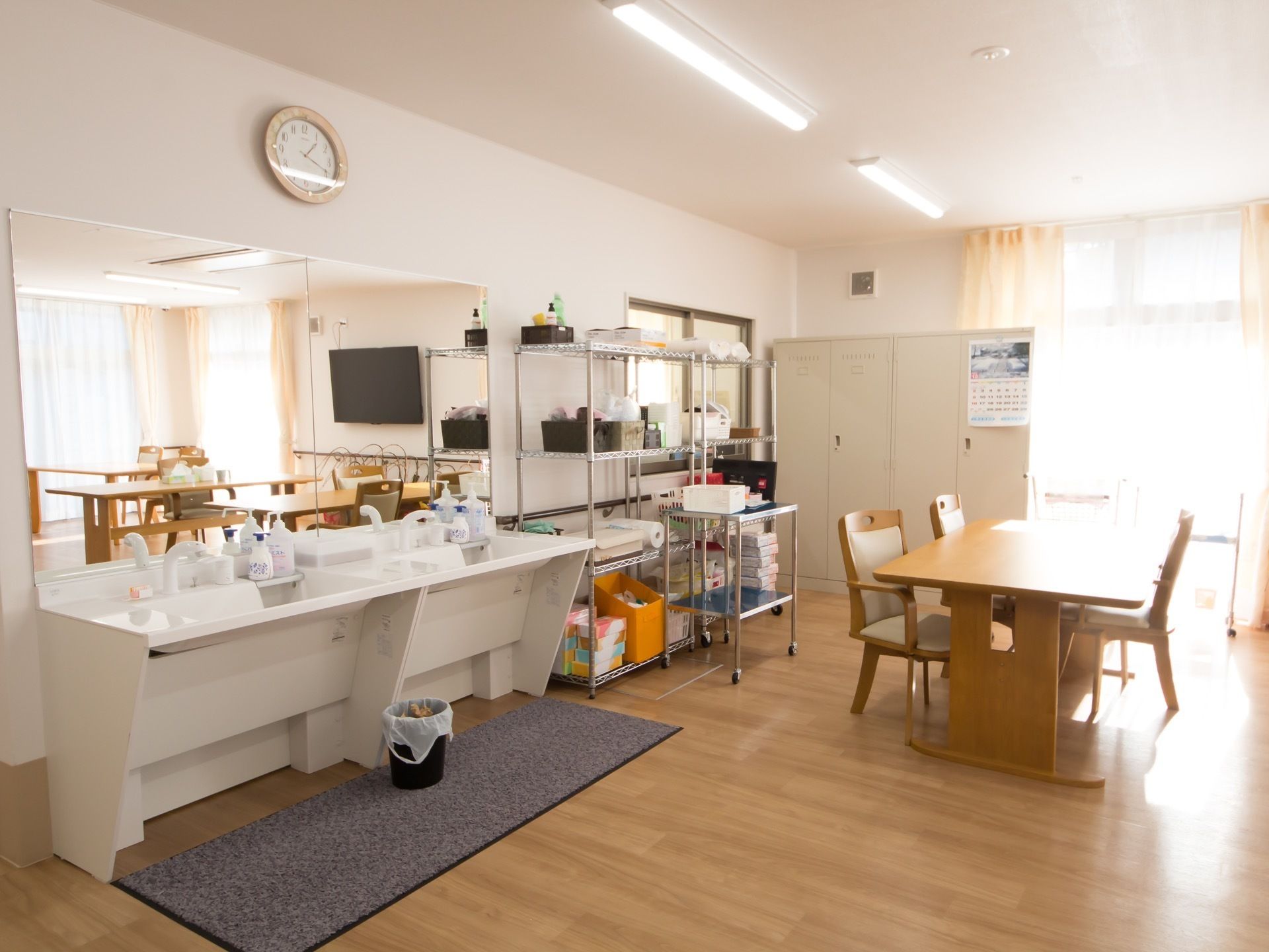 大きな窓から日差しが降り注ぐ、明るい食堂。大きなテーブルと椅子が置かれている。鏡と洗面台があり、壁際には収納棚がある。