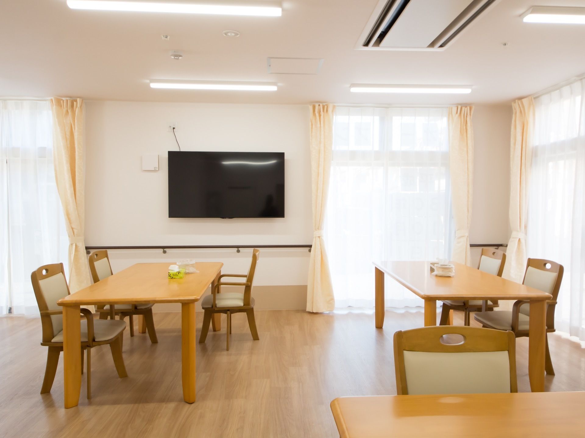 大きな窓から明るい日差しが差し込む食堂。大きなテーブルと椅子が並び、壁には手すりが設置されており、壁掛けのテレビがある。
