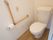 フローリングと白い壁に囲まれたトイレがある。壁には縦横方面に手すりが設置され、非常用ブザーがついている。