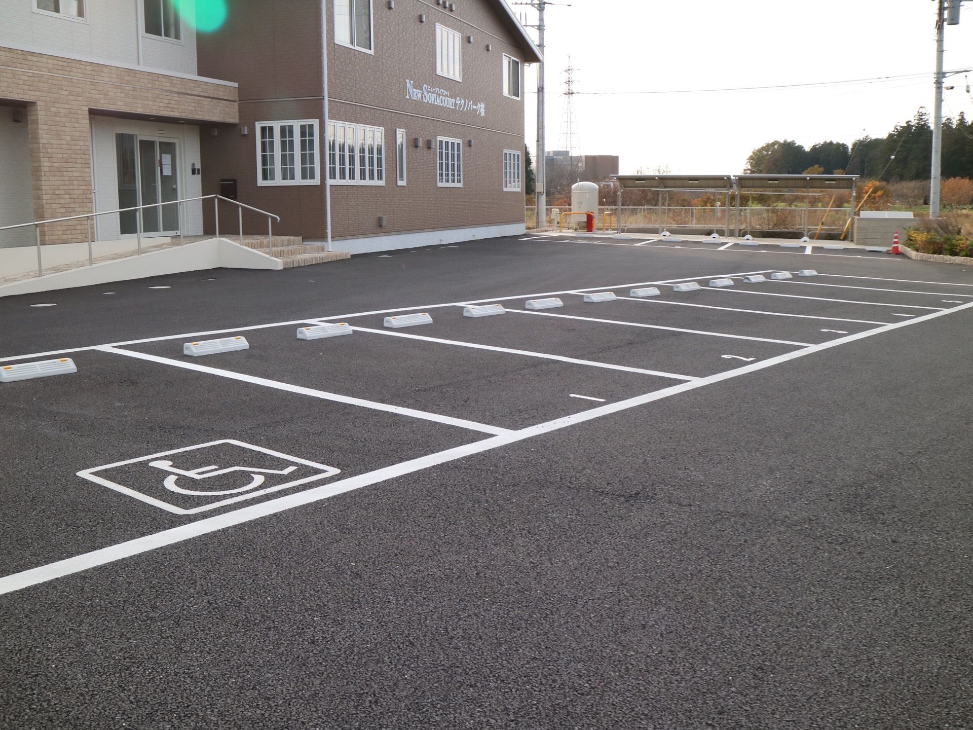 左手奥に２階建ての施設が見えている。その前にはアスファルトで舗装された駐車場が広がっている。車椅子スペースがある。