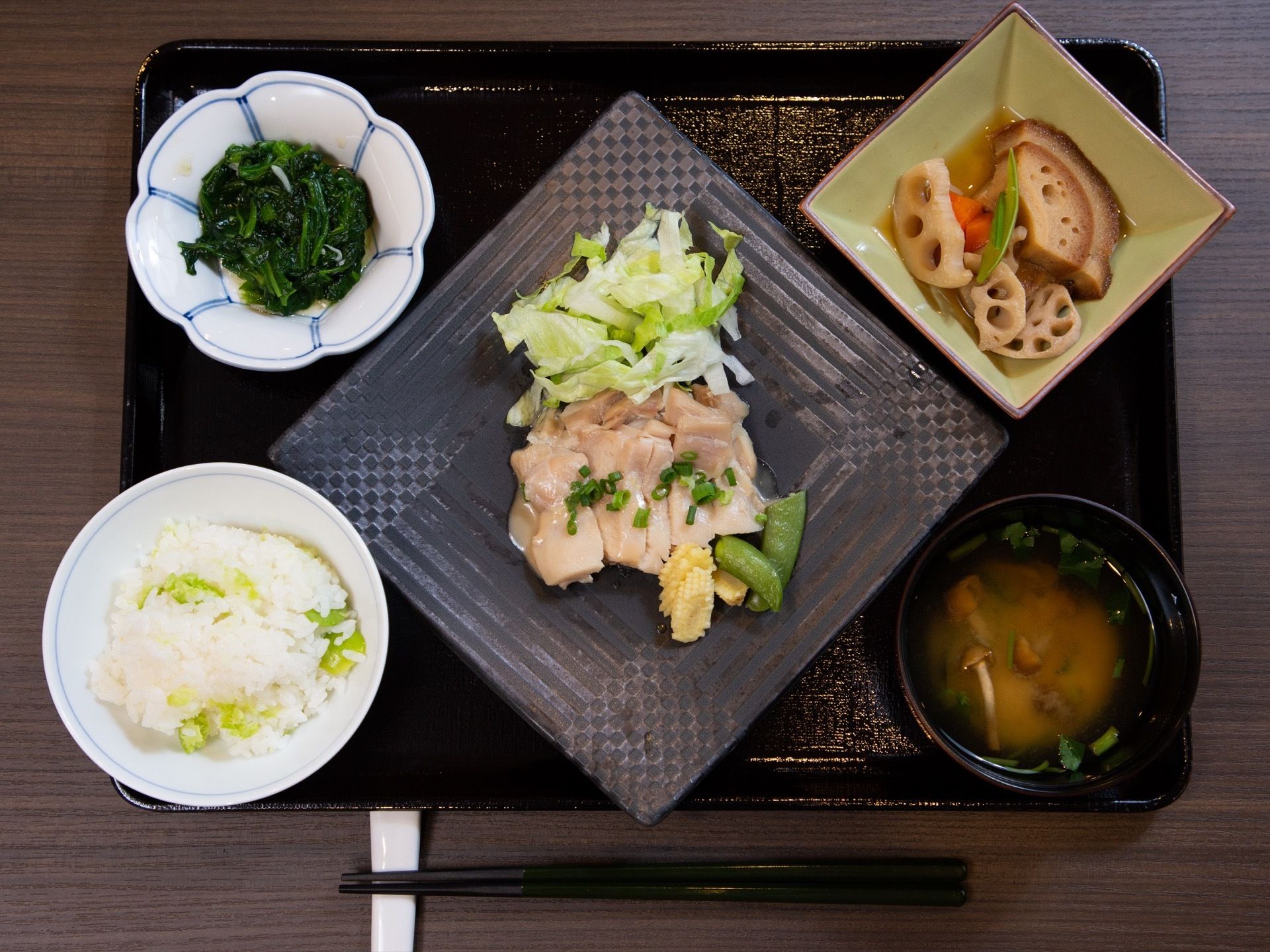 黒いお盆に中央に主菜のお肉のおかず、手前左にご飯、右にお味噌汁、奥左に和え物、右に根菜類の炊き合わせの小鉢がある。