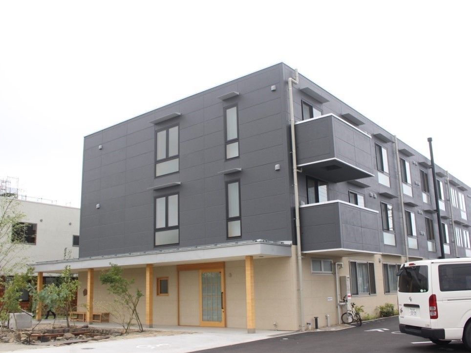 ３階建てのどっしりとした建物は落ち着いた色合い。建物周辺には駐車場があり、緑も植えられている。