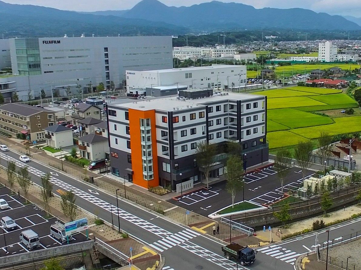 空撮した建物の様子。舗装された車道を臨み、背後には田んぼが広がっている。