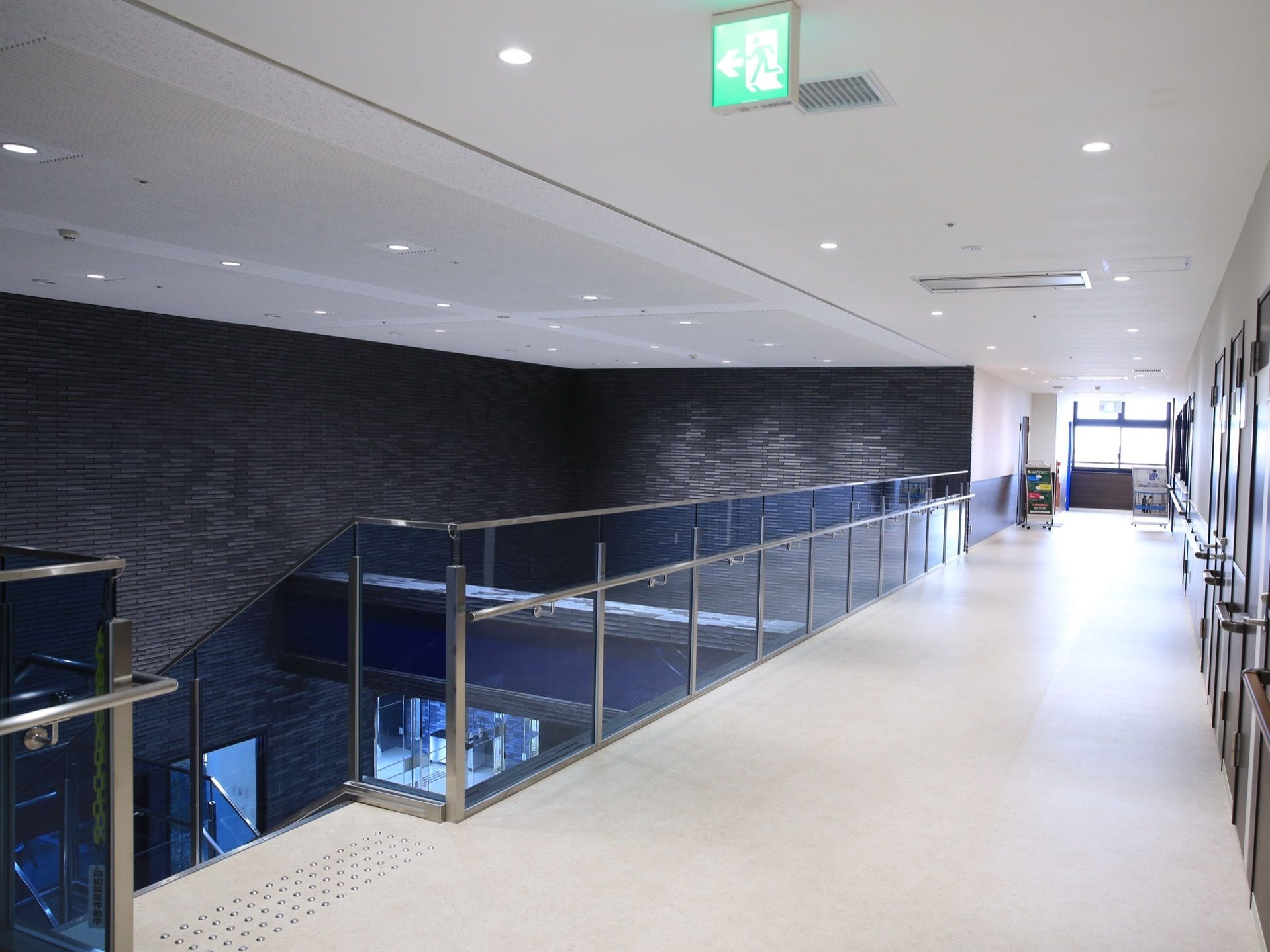 白い床で広めの通路になっている。天井には誘導灯が設置され、左側の階段の前には点字ブロックが付いている。