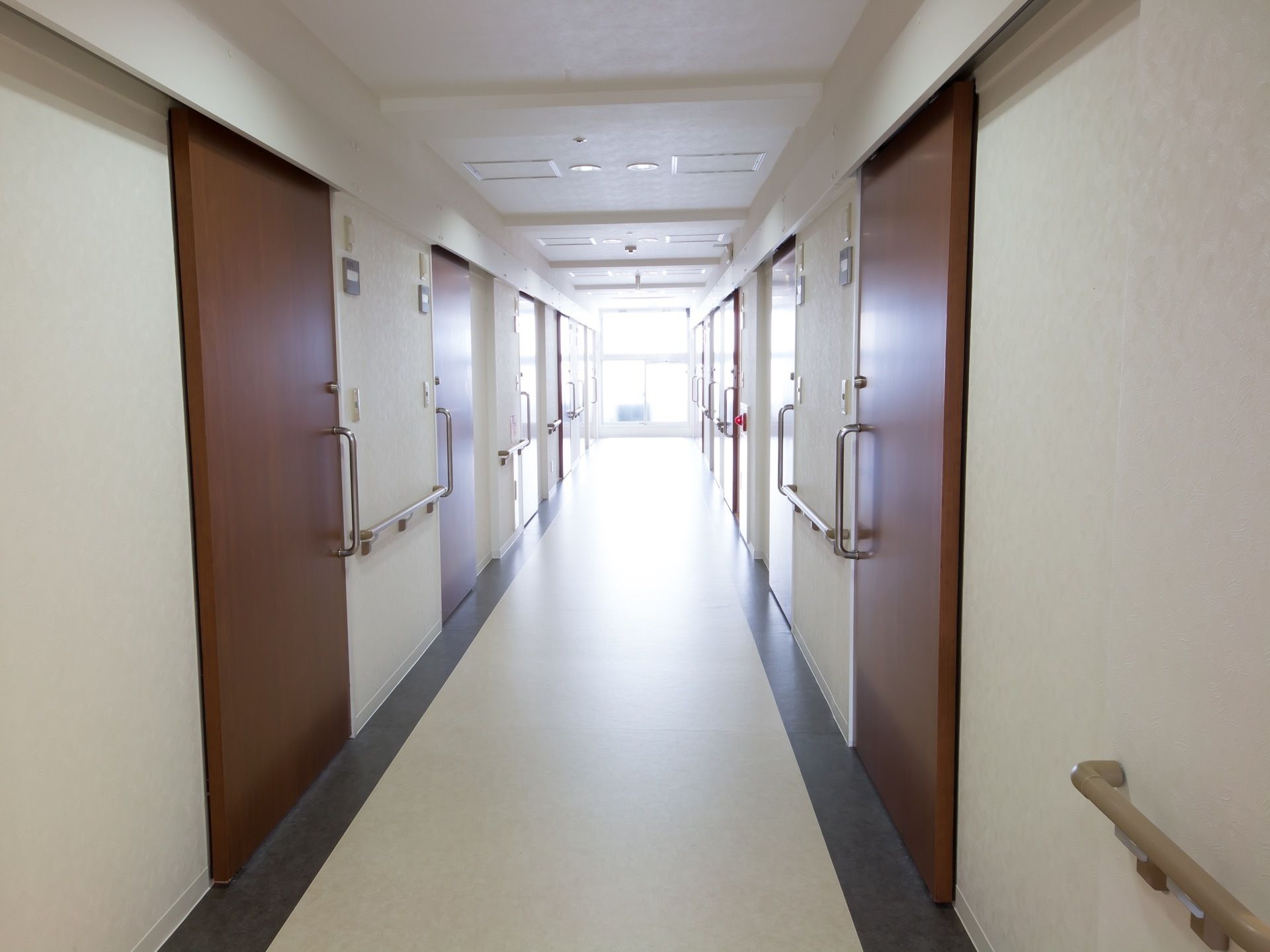 白い床の廊下で両側に引き戸の入口扉が並んでいる。突き当りには窓があり、たくさんの外光が差し込んでいる。