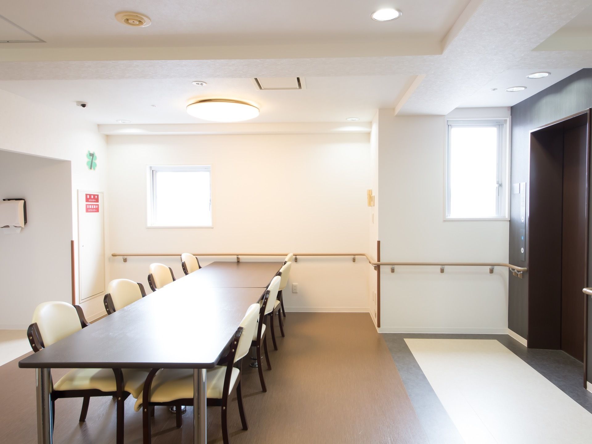 右手に黒いエレベーターの扉がある。４人掛け用のテーブルセットが縦に２セット並べて置かれている。窓が２か所に開いている。