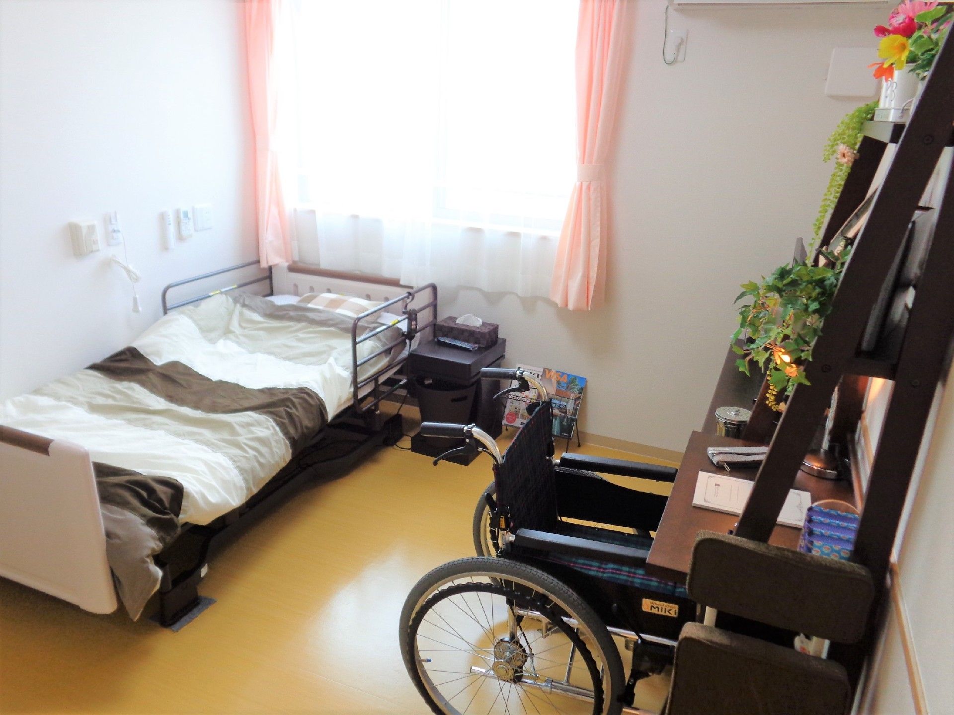 布団がセットされたベッドが左側にあり、収納家具や車いすも置かれた部屋。