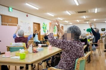 車椅子に乗って手を振る高齢女性と介助をする男性ヘルパー
