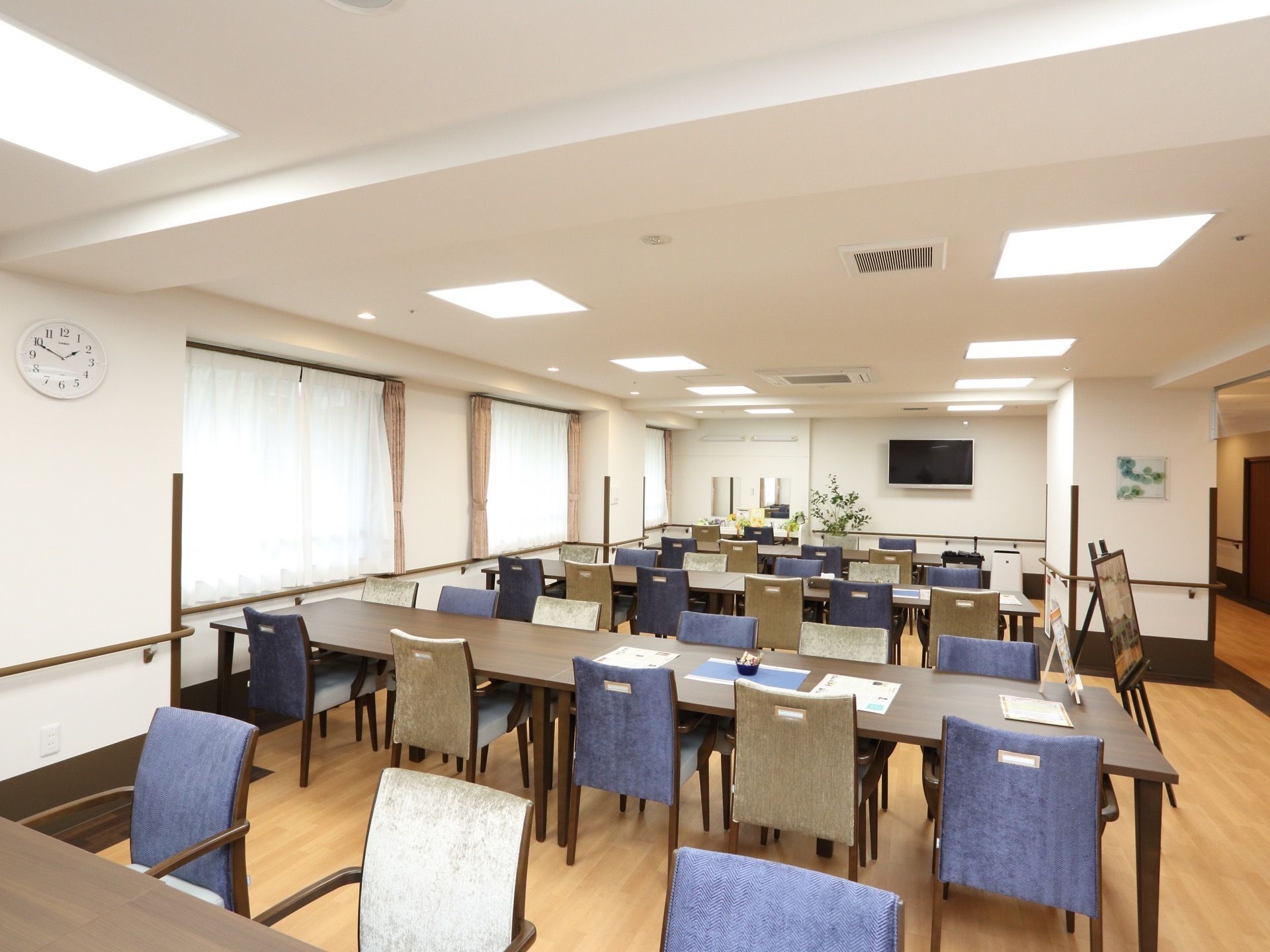 天井、壁が白色の広い部屋である。横長のテーブルに背もたれがベージュ色と濃いブルー系色の椅子がきれいに交互に並べられ、数組セットされている。