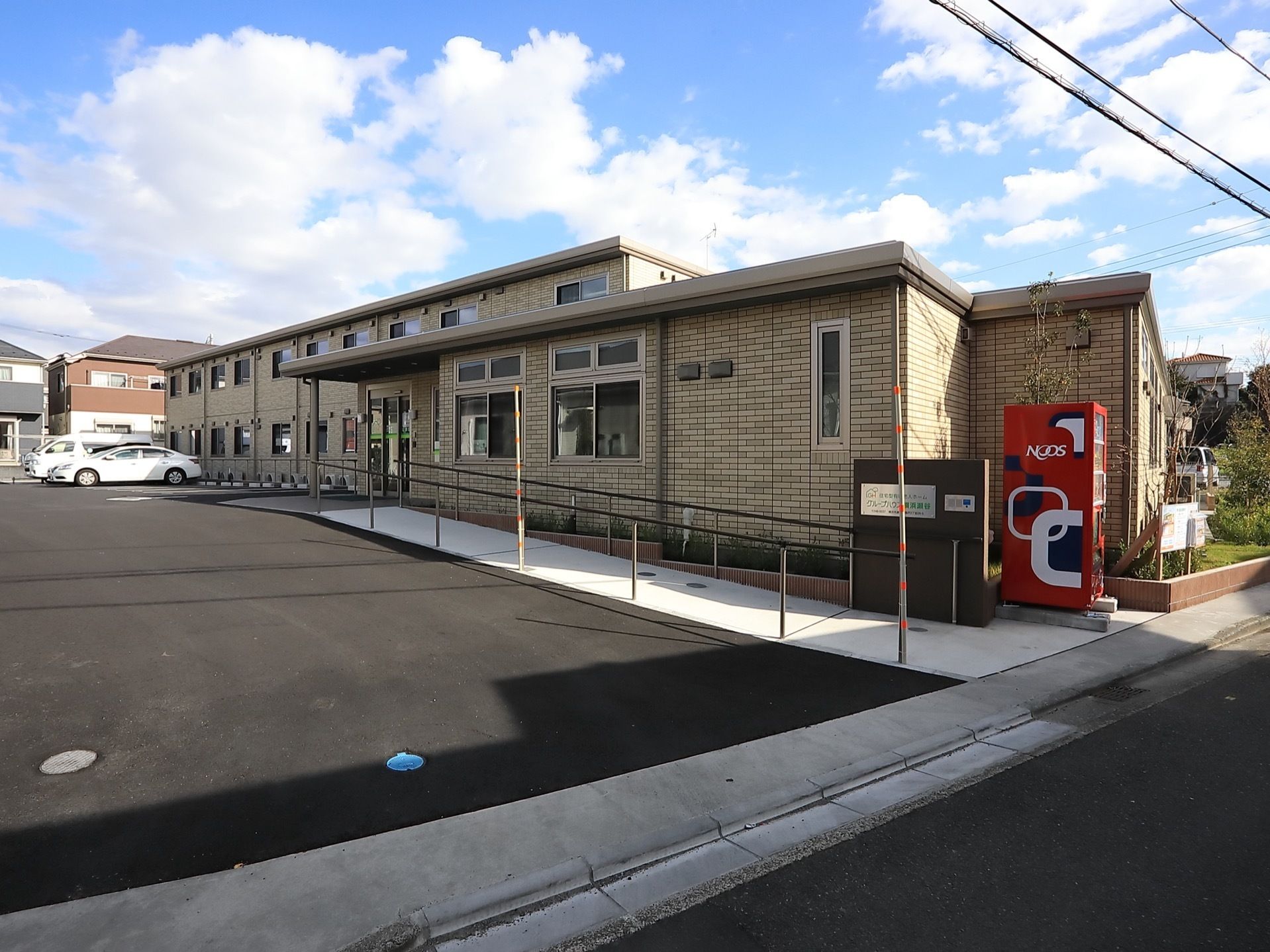 道路から奥行のある敷地に建つ２階建ての施設である。玄関部分は平屋建てになっている。道路側に自動販売機がある。