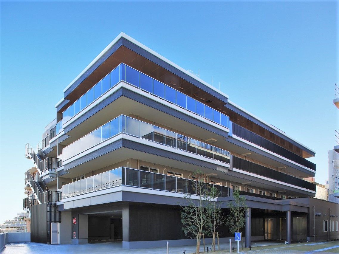 4階建ての大きな建物はグレーを基調とし、1階部分には駐車スペース、建物前には季節の木や花が植えられたスペースがある。