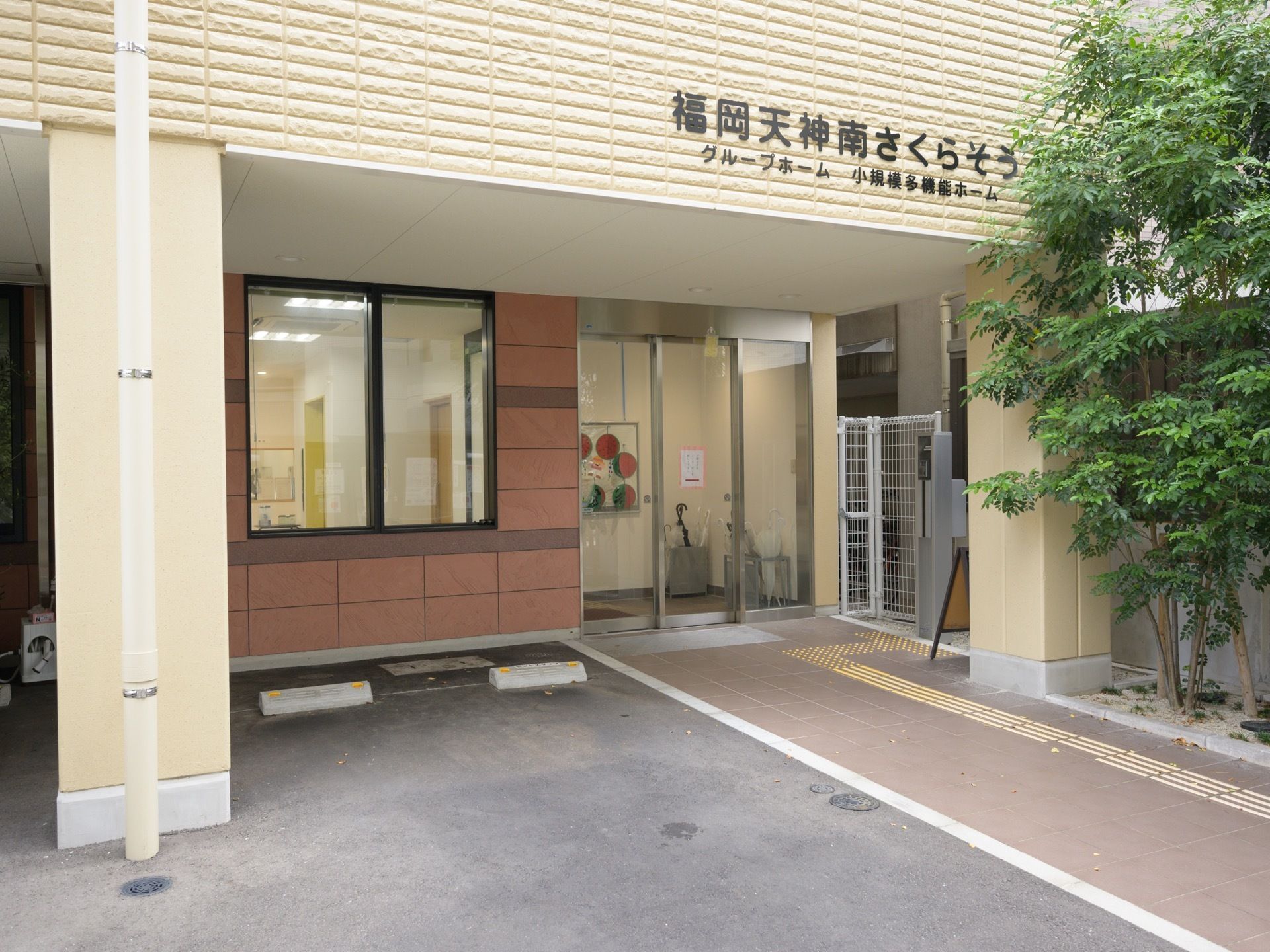 ベージュ調の施設外観となっており、外壁には施設名が記されている。駐車場が設けられており、通路には点字ブロックがある。