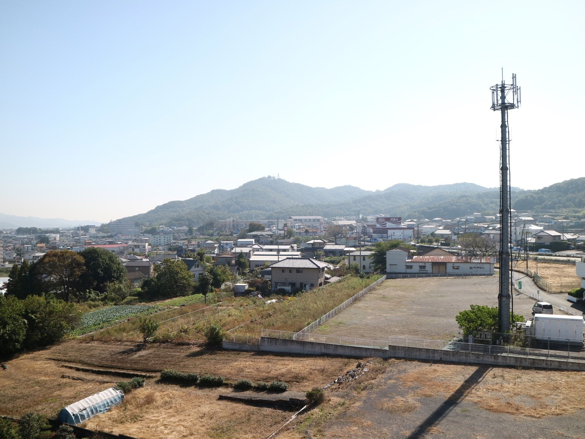 施設内から見える眼下の風景。目の前の空き地脇に高い鉄塔が建っている。横にハウスなどのある畑が広がっている。その奥に住宅街と山並がある。