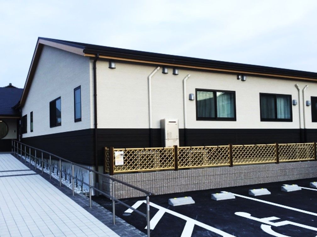 入り口にスロープがあり、濃茶とベージュの外壁で三角屋根の平屋のグループホーム。
