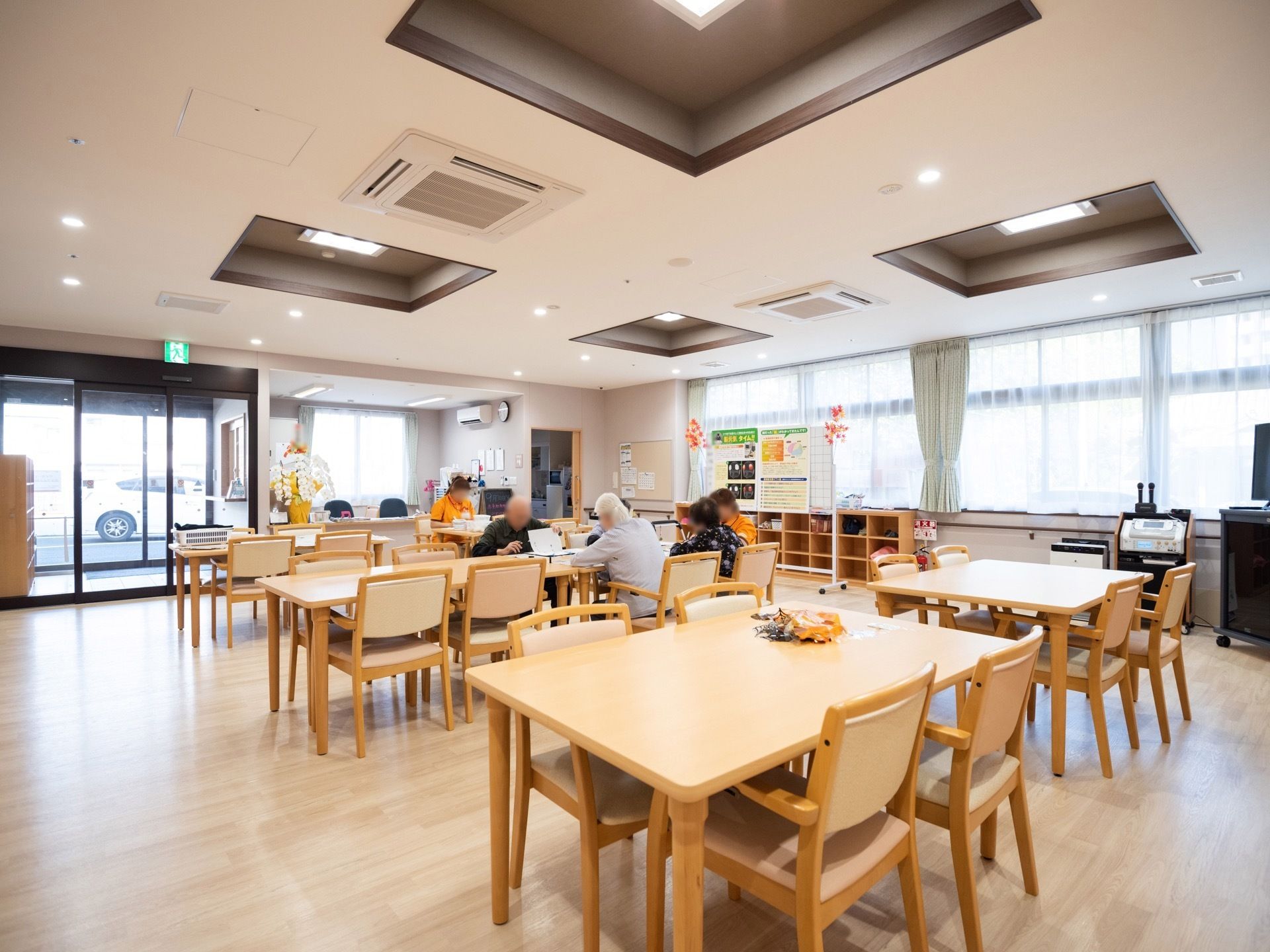 スタッフと入居者様がテーブル席に座って作業を行っている。窓の近くに置かれたホワイトボードにポスターが貼られている。