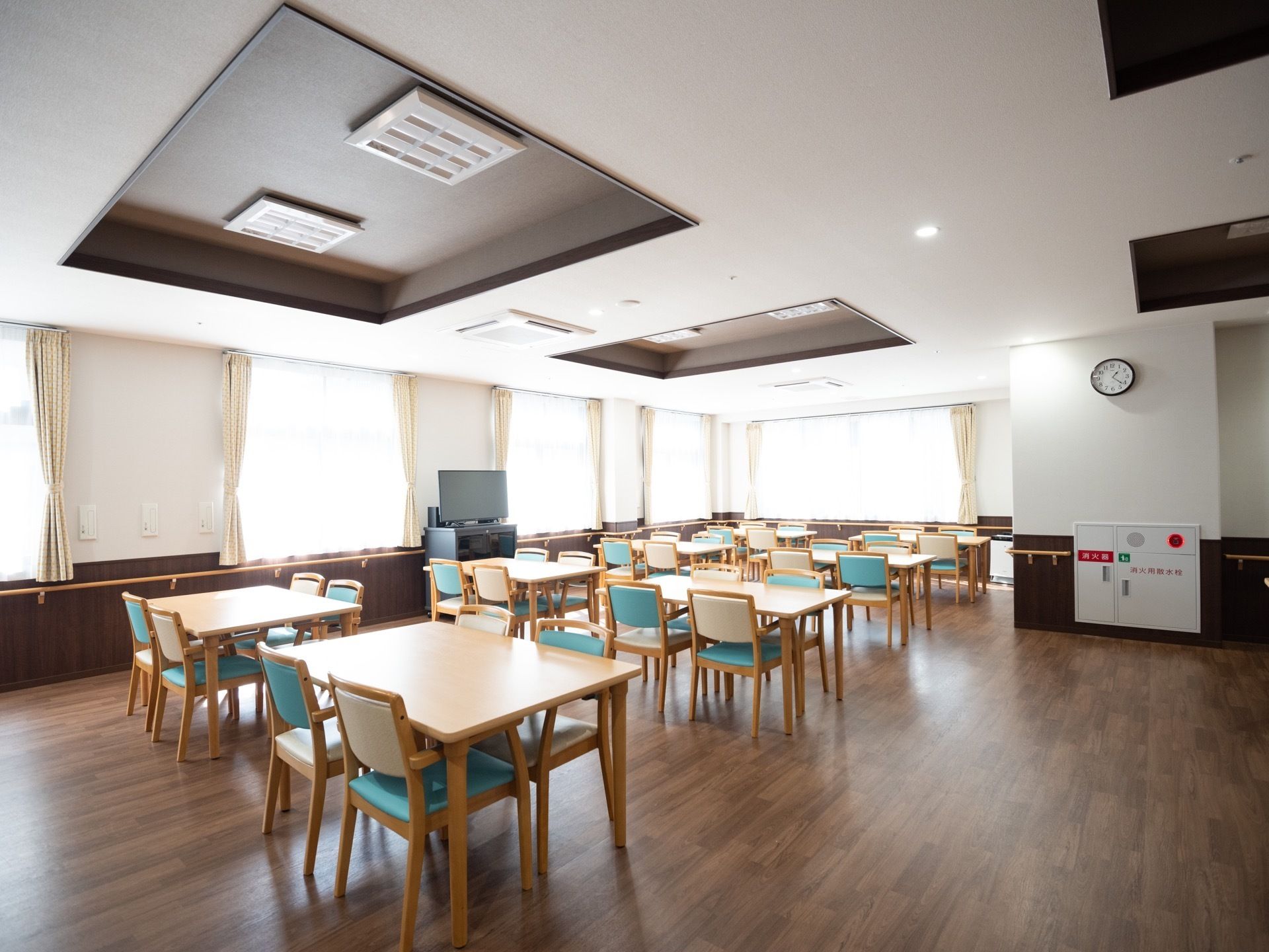 食堂にある掘り込まれた天井に照明器具が設置されている。窓と窓の間の壁の前にある台の上にテレビが置かれている。