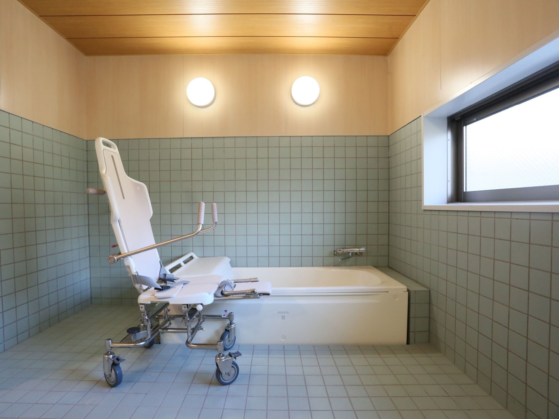 水色のタイルが床と壁に貼られた浴室である。横長の窓があり、窓に向かって浴室用の車椅子が１台置かれている。