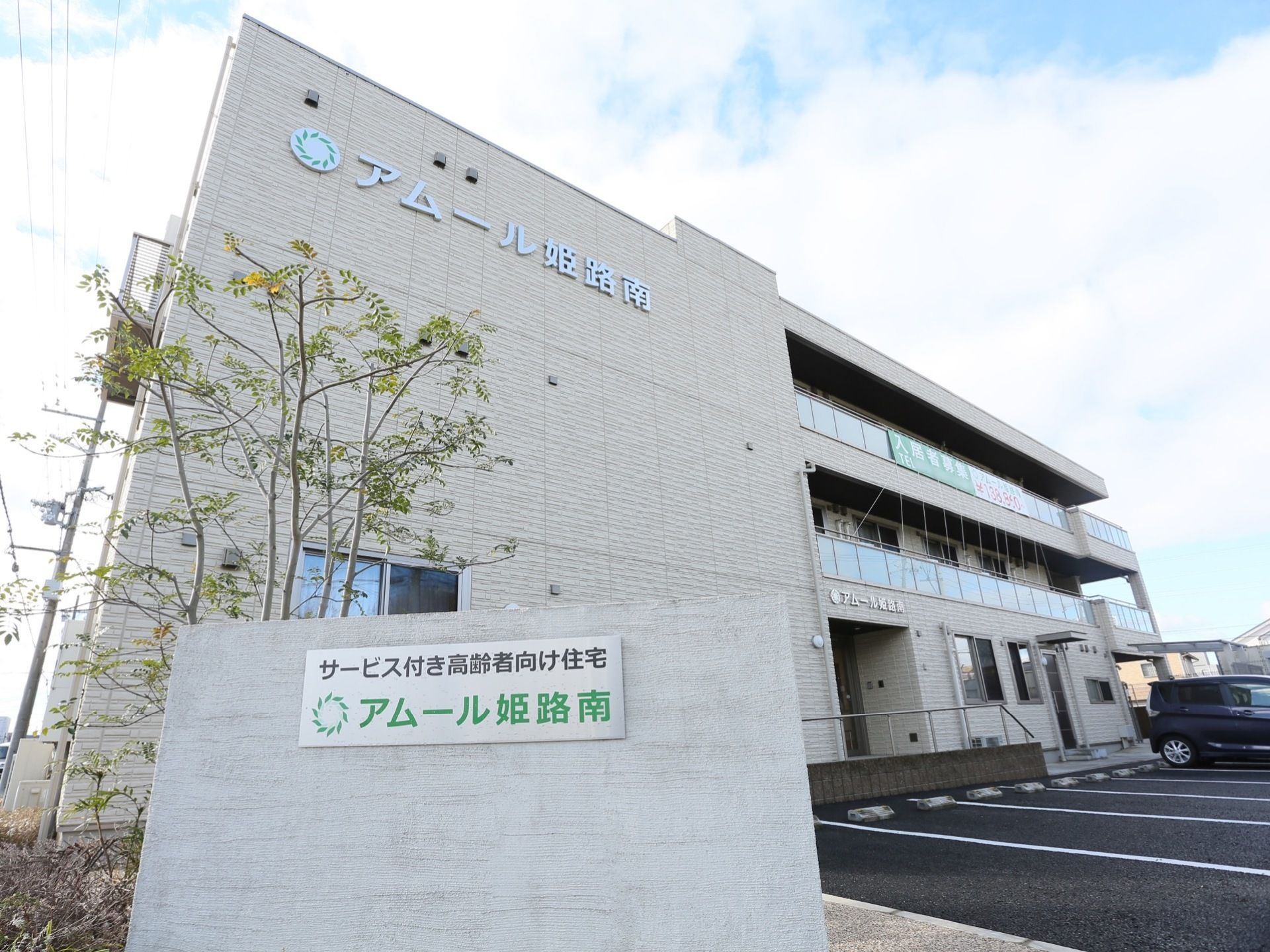 ３階建てのグレーの施設である。施設の前は駐車場になっており、奥に車が１台停まっている。入口に看板が置かれている。