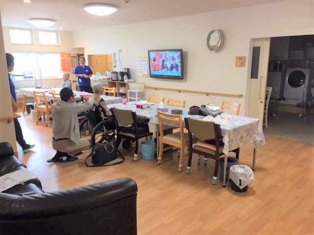 明るく広い共有スペース。車いすの入居者がダイニングテーブルにつき、スタッフが数名フロアで働いている。