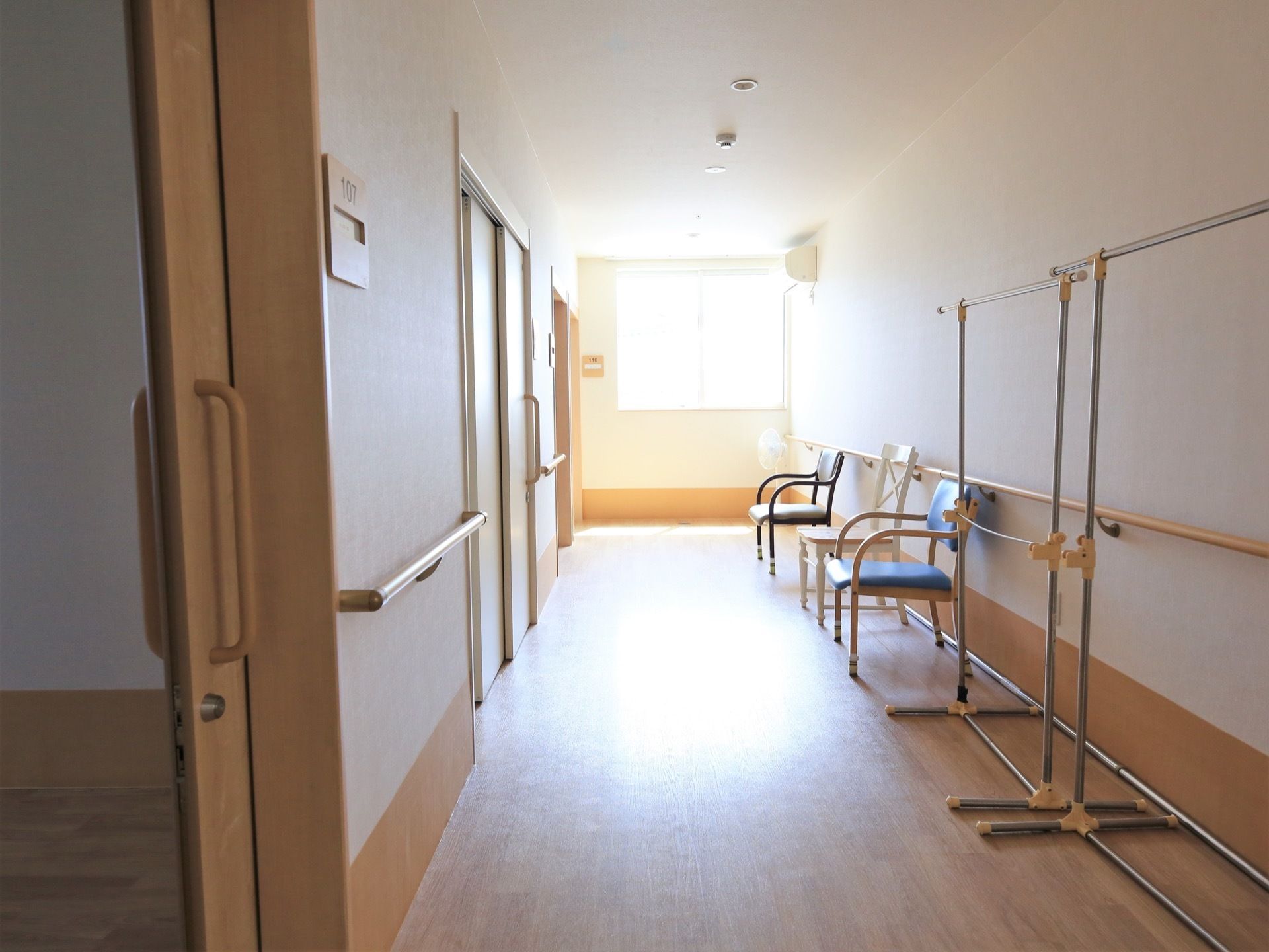 つきあたりにある窓からの採光で明るい廊下である。壁際に背の高いパイプハンガーが２つと椅子が３脚並んで置かれている。