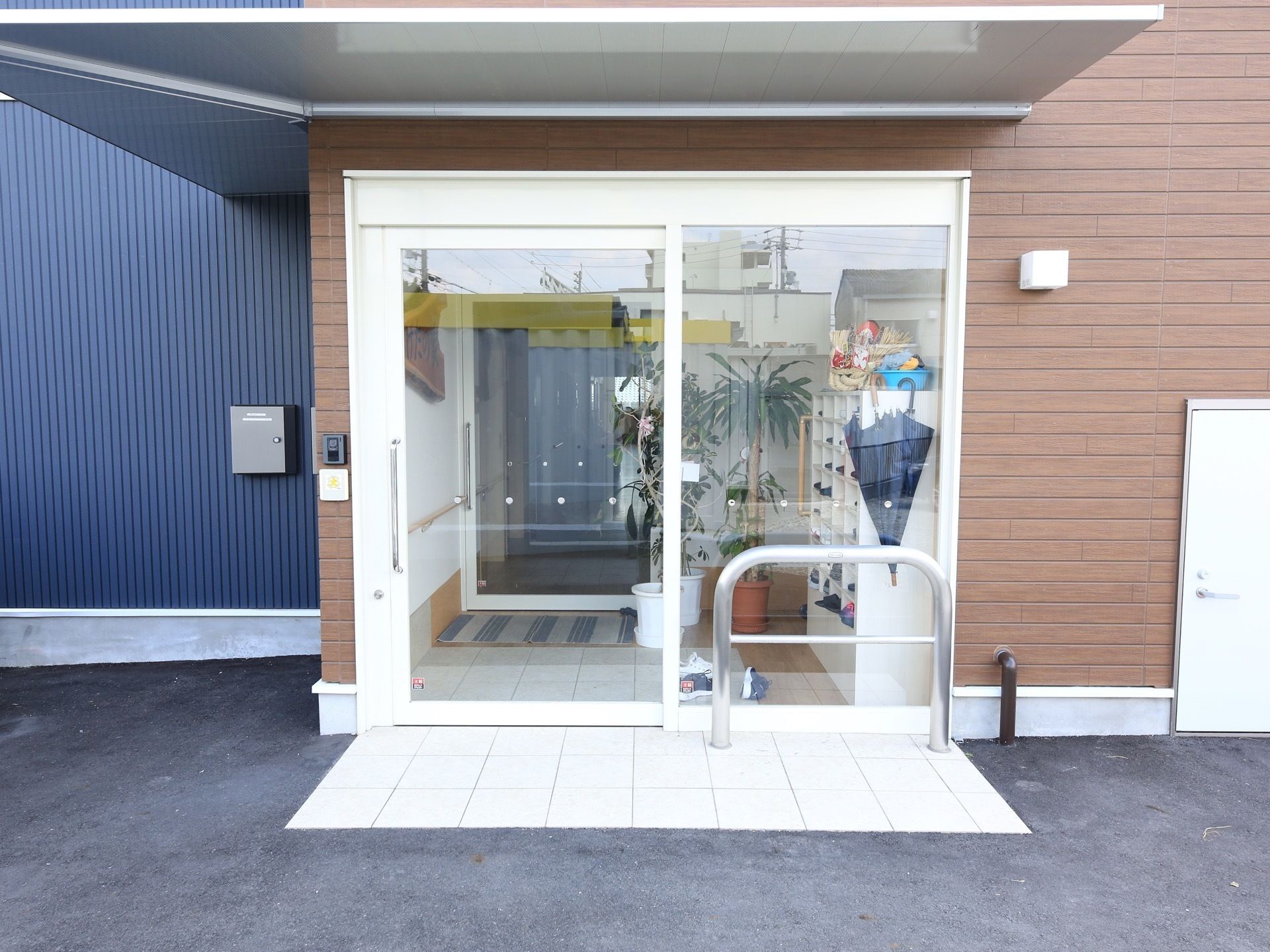 玄関の扉は、縦方向の大きな取っ手がついた白色枠のガラスである。左側の外壁にインターホンが設置されている。