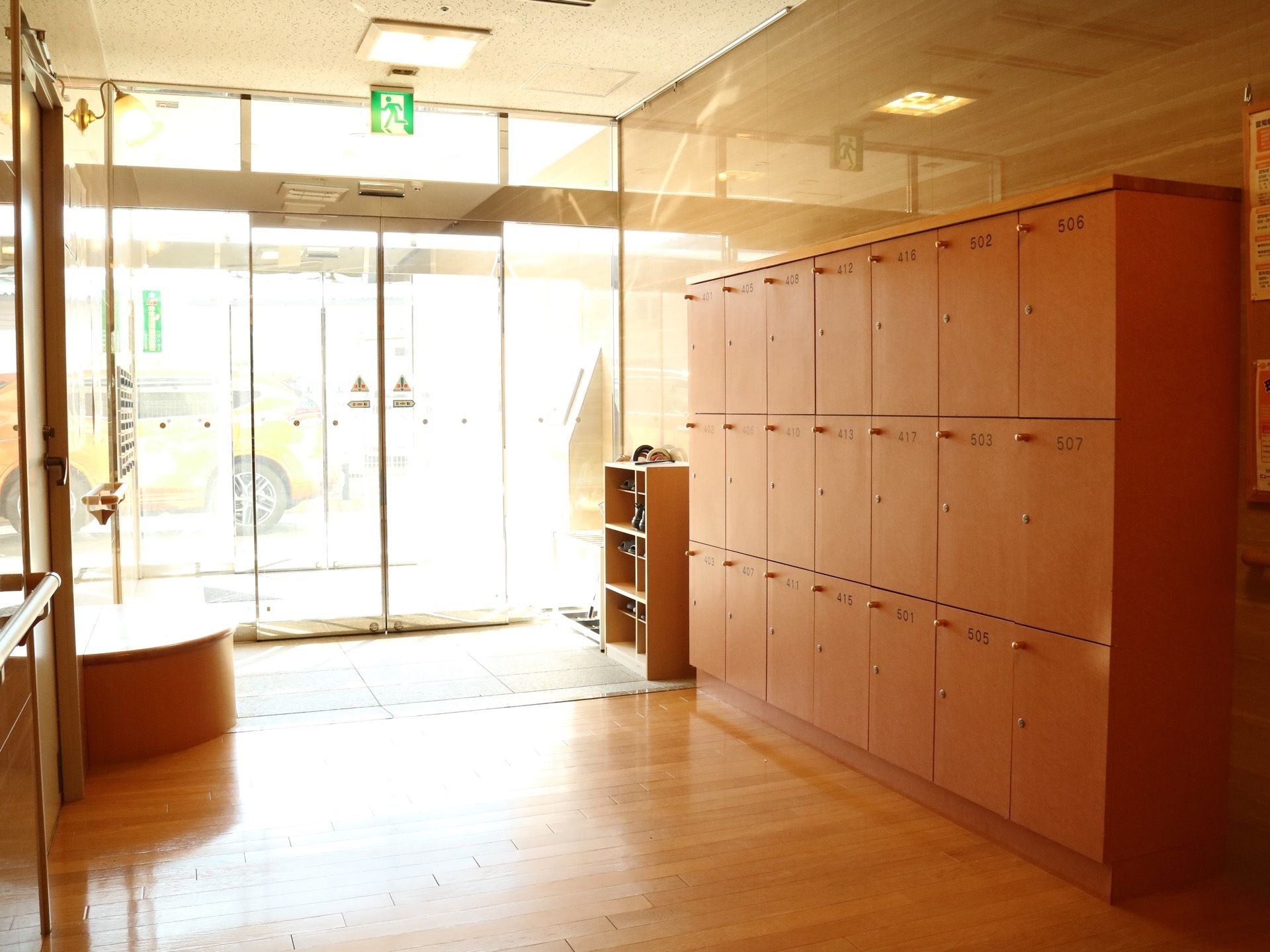 扉付きの大きな木製の靴箱が設置されているエントランスで、向かい側に椅子が用意されている。ドアの上には、非常口の看板が表示されている。
