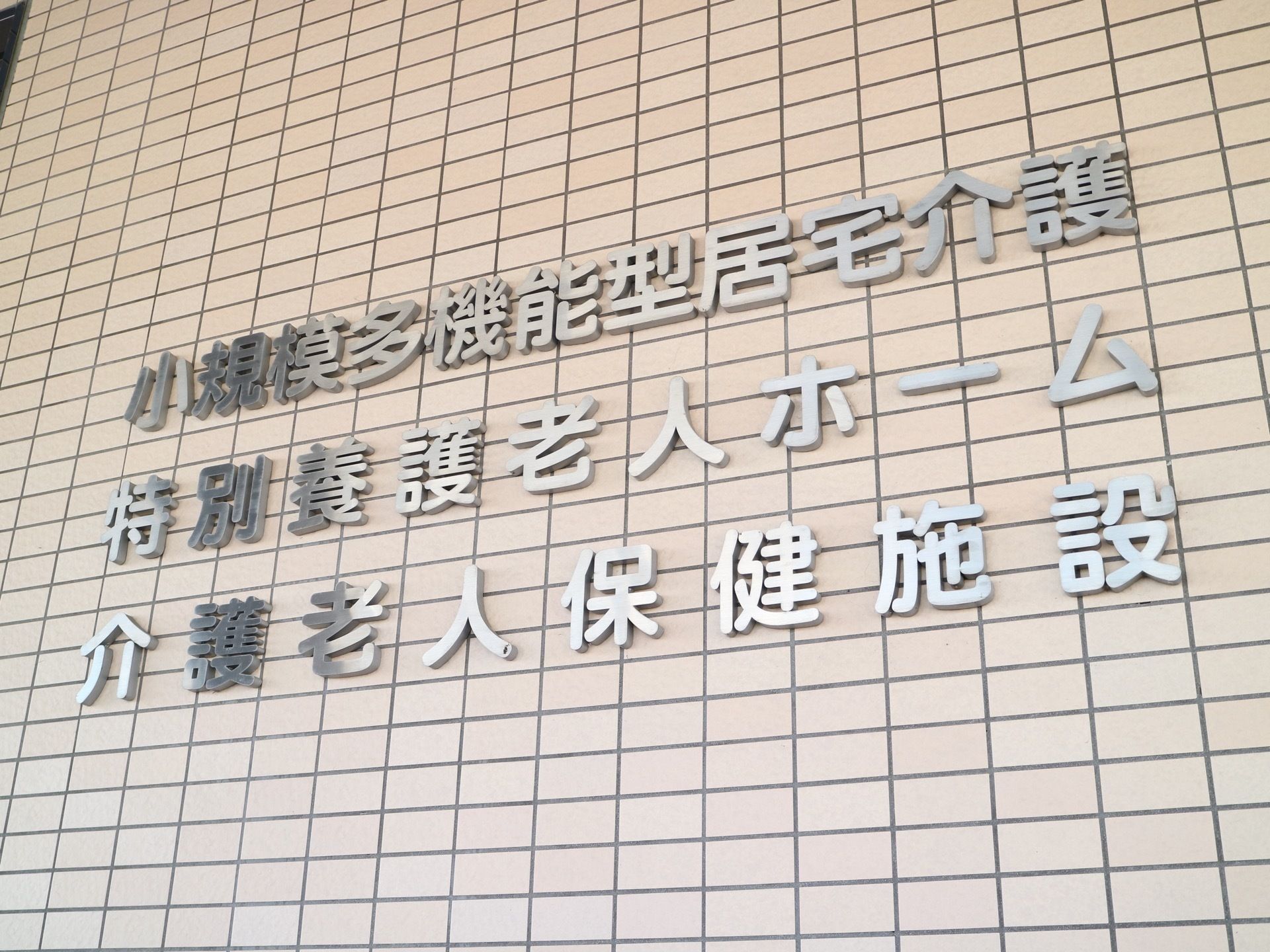 複数の事業所が併設されているグループホーム。白を基調としたタイル張りの外壁に、銀色の文字で施設名が書かれている。