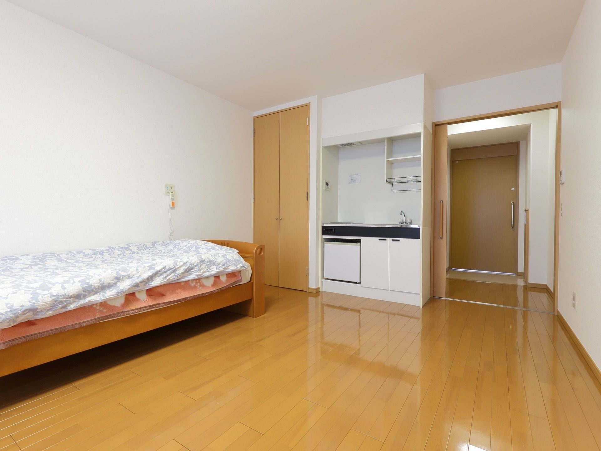 フローリングの居室。IHヒーター付のミニキッチン、収納、ベッドがある。壁には緊急時の呼出しボタンが設置されている。
