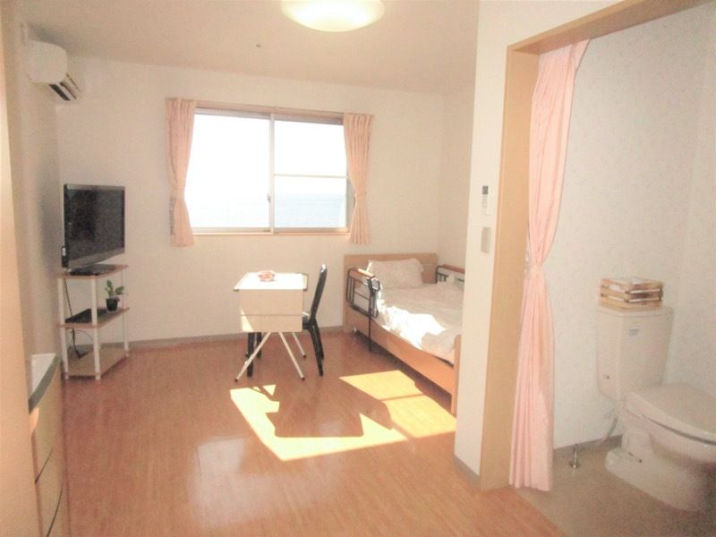カーテンで開け閉めするタイプのトイレや寝具付きのベッドが付いたフローリングの部屋。