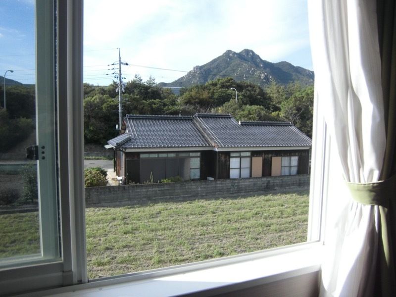 カーテン付きの窓を開けて周辺を見た様子。平屋建ての民家があり、田畑が広がっている。