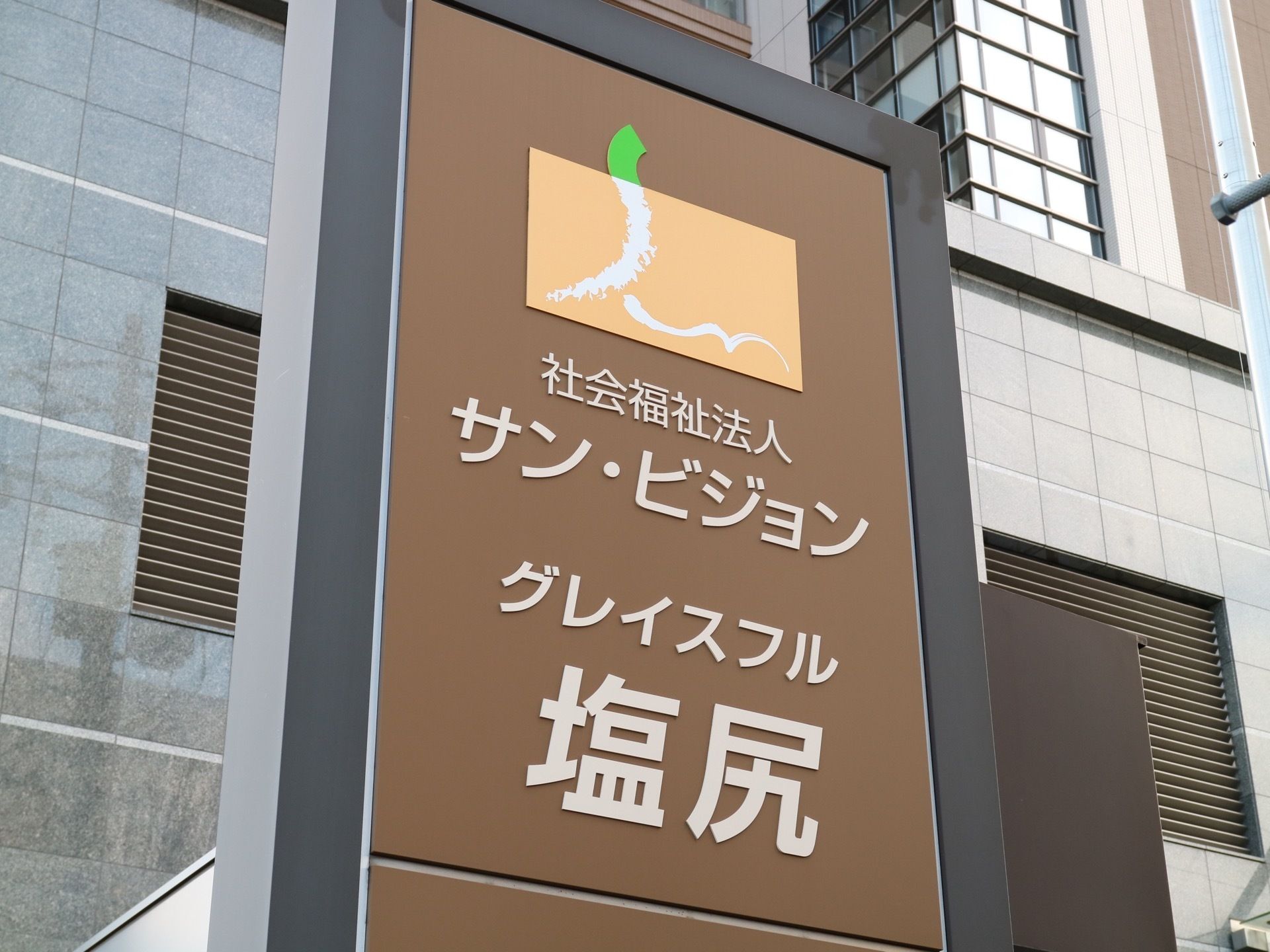 施設名の書かれた茶色の看板が立っている。後ろにはグレーの外壁の建物があり、上の階には窓が多く付いている。