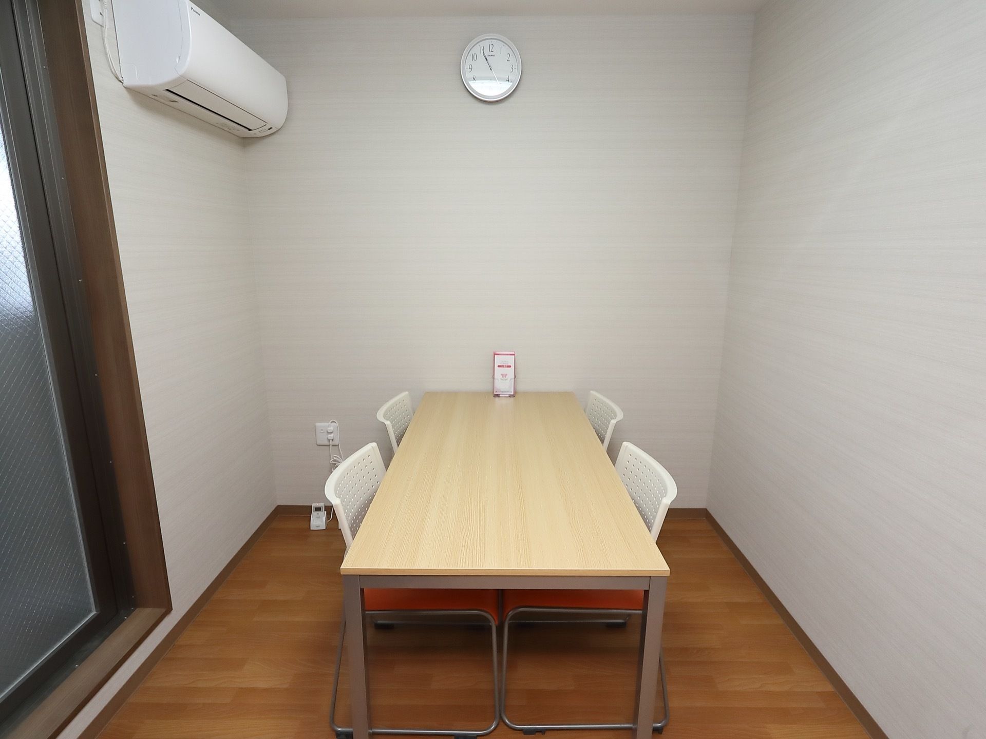 窓のあるフローリングの部屋である。ナチュラルな色合いの長方形のテーブルと白い背もたれの椅子が４脚設置されている。