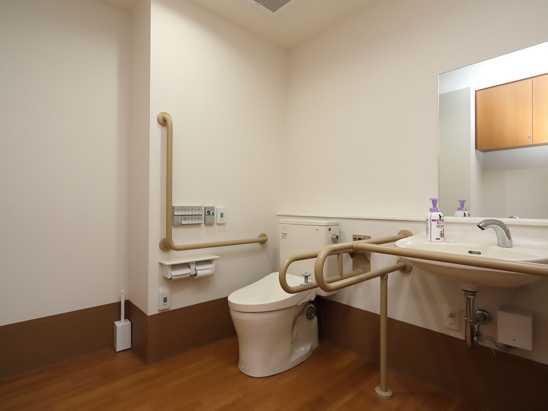 広々としたフローリングのスペースに、トイレと鏡付きの洗面台が並んで設置されている。手すりが各所に取りつけられている。
