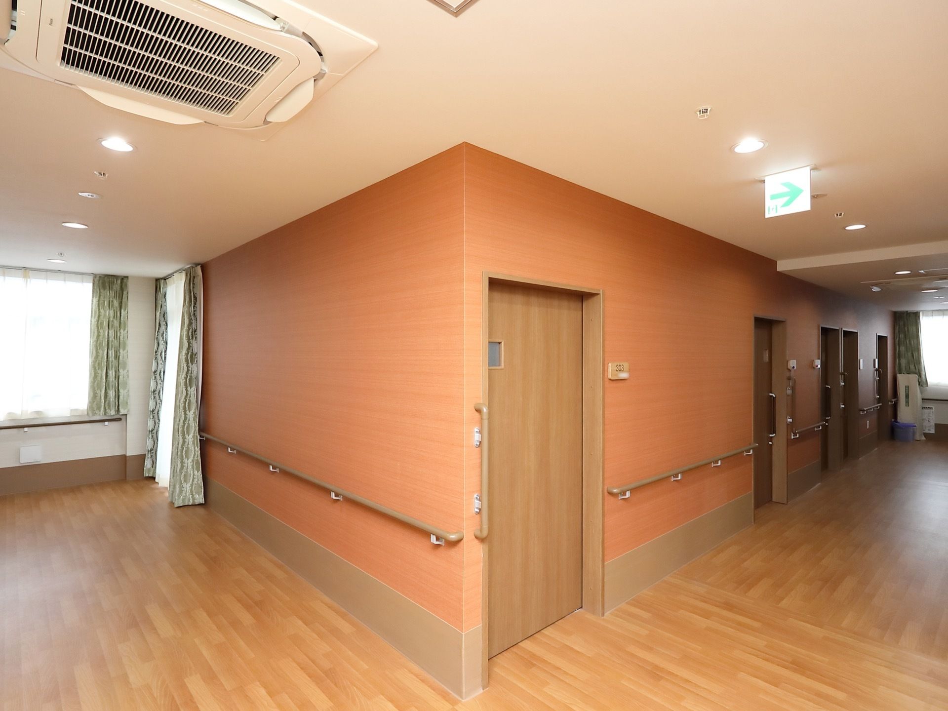 通路の曲がり角から居室ドアが並んでいる。周囲の壁はオレンジ色っぽい木目調で、手すりが取りつけられている。
